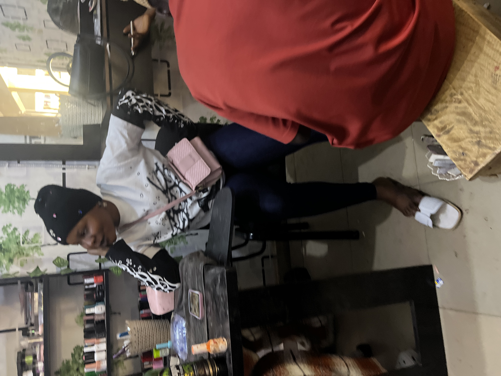 The image shows an indoor scene with three individuals. A woman wearing a black beanie, white shirt with black patterned sleeves, blue jeans, and white sandals sits on a black chair, a pink purse slung over her shoulder. Her head is resting on her hand. In the background, there's a person in red attire, partially visible and cropped out of the image. Another person is partially visible in the top-left.
To the left is a shelf containing numerous bottles, possibly nail polish. The setting appears to be a beauty salon or similar establishment, possibly located in Jos, Nigeria, given the contextual request. The floor is a light color. Additional furniture is visible under the woman's chair.
