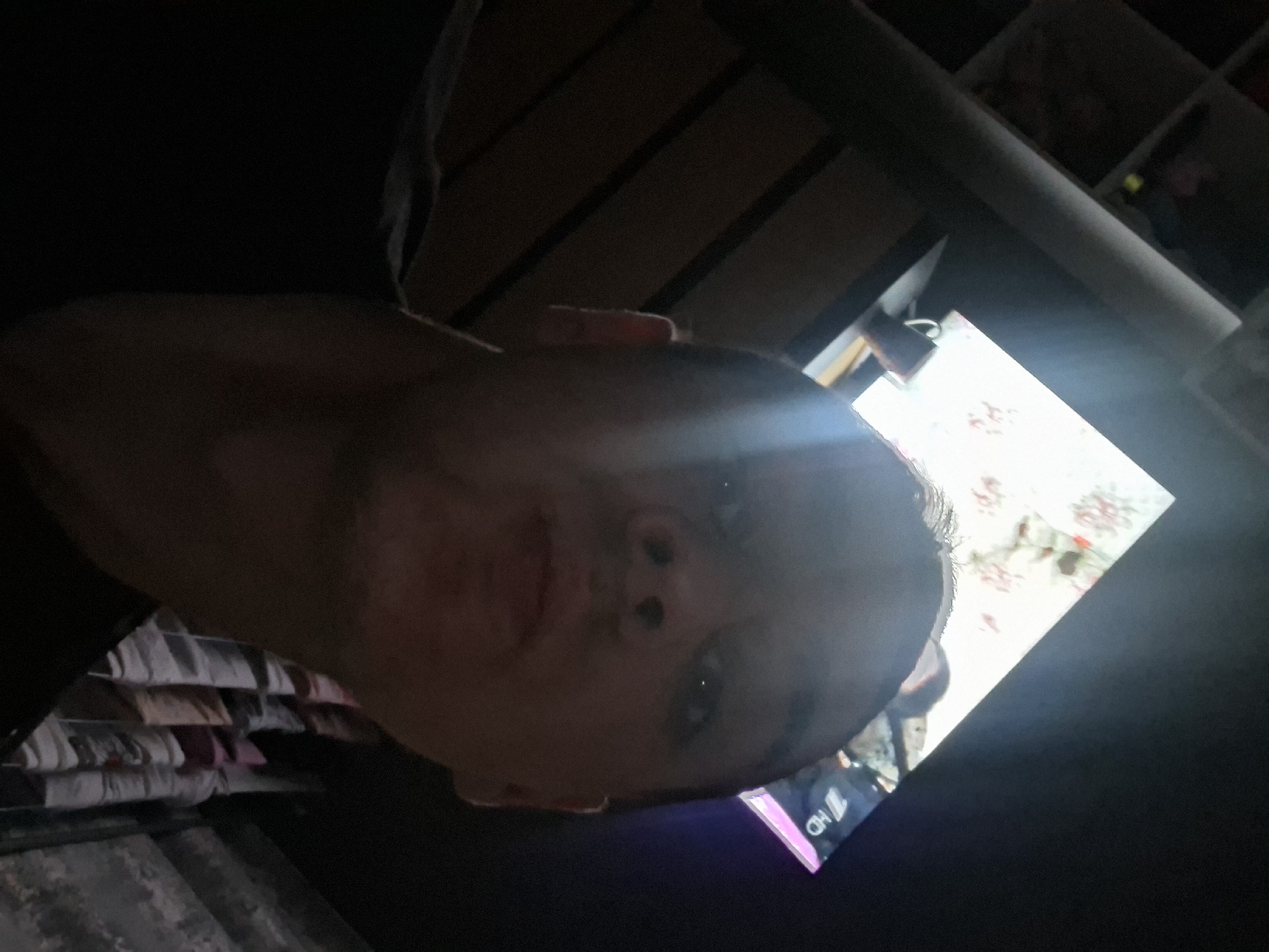 A person's face is centrally located in the image, illuminated by a bright light source from above. The person appears to be looking directly at the camera. The background is dark and obscured, with hints of shelving and miscellaneous items. A rectangular shape is slightly visible on the right side of the image. The location appears to be inside a building. City information is unavailable.