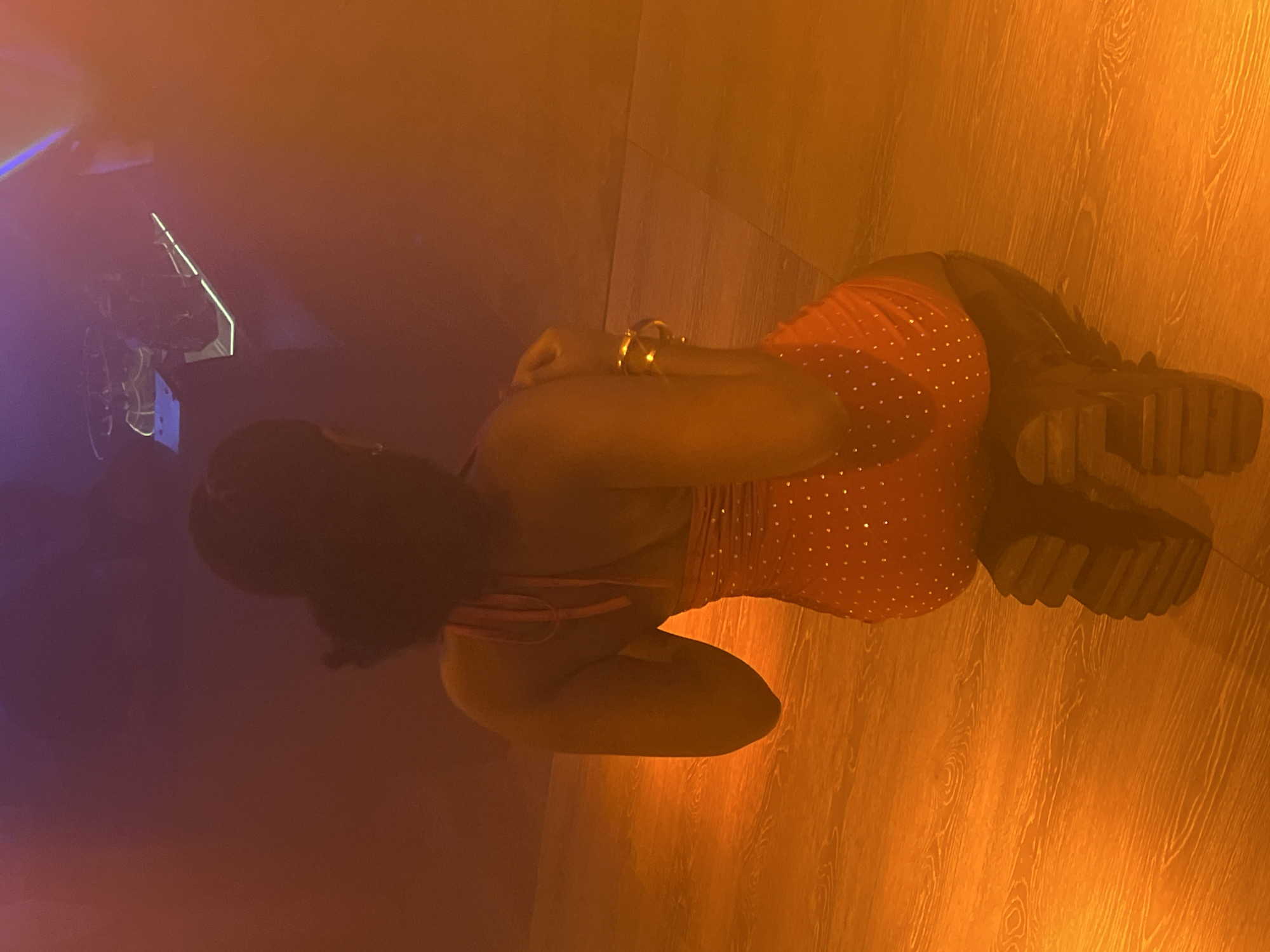 A woman is lying on a wooden floor, wearing an orange top with silver studs and black boots. The lighting is orange, suggesting indoor lighting. A machine is visible in the background under blue light. The location is consistent with a nightclub or bar in Howayi, Nigeria.