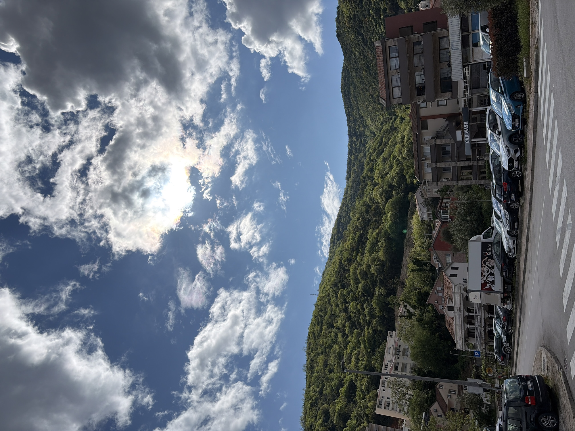The image shows a street scene in Budva, Montenegro, featuring a view towards a tree-covered mountain with buildings nestled along its lower slopes. A multi-story building stands on the right edge of the frame, adjacent to a row of parked cars. The cars are parked along the edge of a street, marked by a zebra crossing. The sky above is partly cloudy with a bright sun obscured by the clouds. The overall scene conveys a sense of being in an urban environment with natural surroundings.