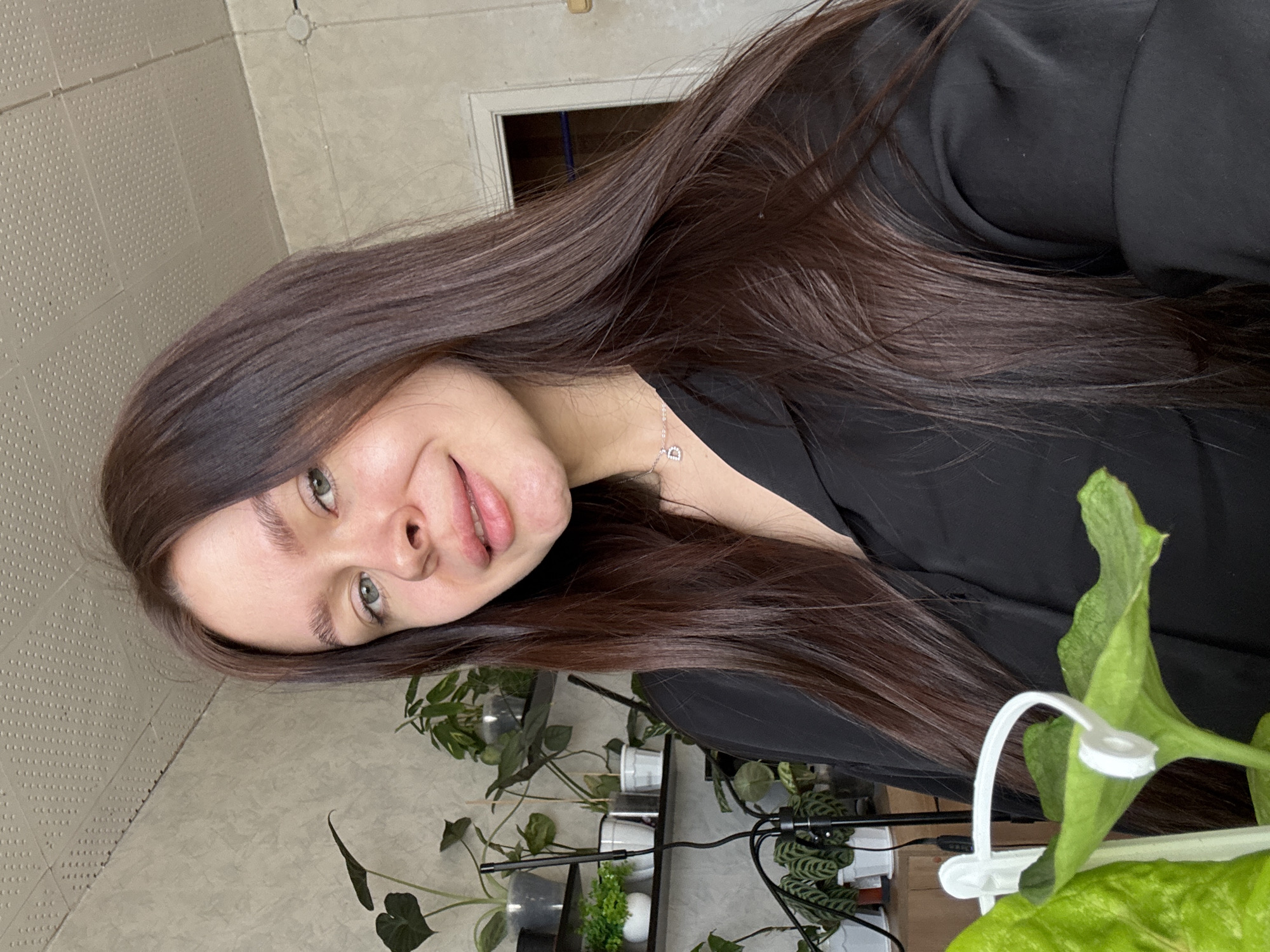 A young woman with long, dark brown hair is positioned in the foreground of the image, facing the camera at an angle. She has fair skin and her eyes are green. She is wearing a black top and a delicate silver necklace. Behind her, a collection of potted plants is visible on a shelf, suggesting an indoor environment. The background wall has a textured pattern. The presence of a plant in the immediate foreground partially obscures the view. There is nothing in the image to indicate this is from Dubrova, Russia.
