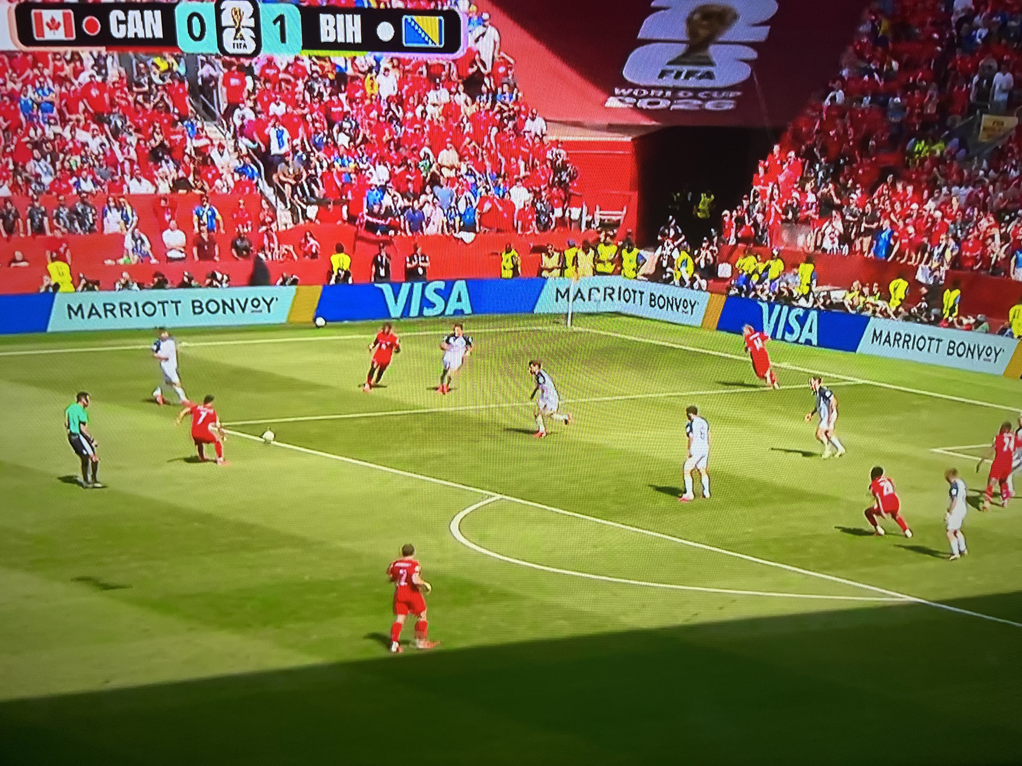 The image depicts a soccer match on a vibrant green field with white boundary lines. Spectators fill the stadium seating, predominantly in red. A digital scoreboard at the top displays "CAN 0 1 BIH" with national flags of Canada and Bosnia and Herzegovina. Prominently displayed banners along the field's edge read "MARRIOTT BONVOY" and "VISA."
Several soccer players in red and white jerseys are visible on the field. One player in a red jersey with the number 7 is dribbling the ball, while others are positioned around the field, some in motion, suggesting active gameplay. The referee, in a black and white striped shirt, stands to the left, observing the play. The overall environment is that of a professional outdoor soccer stadium during a daytime match. The city and specific location are unknown.
