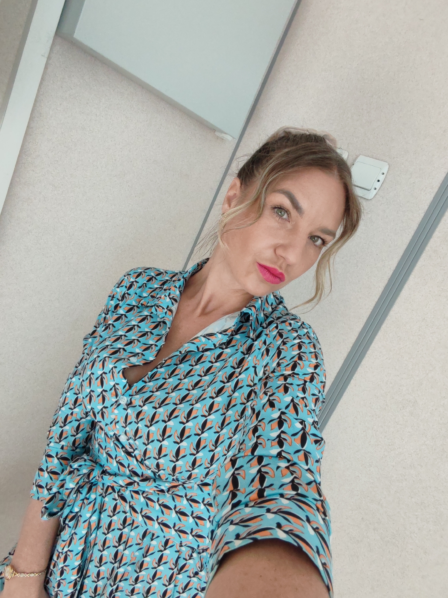 A woman is posing for a selfie. She has blonde hair pulled back with some strands framing her face. She is wearing a blue patterned dress with black and orange accents and pink lipstick. The background consists of a plain wall with a gray trim and a light fixture. The location appears to be indoors, possibly a restroom or office space. The city and specific location within the city are Unknown, Unknown.