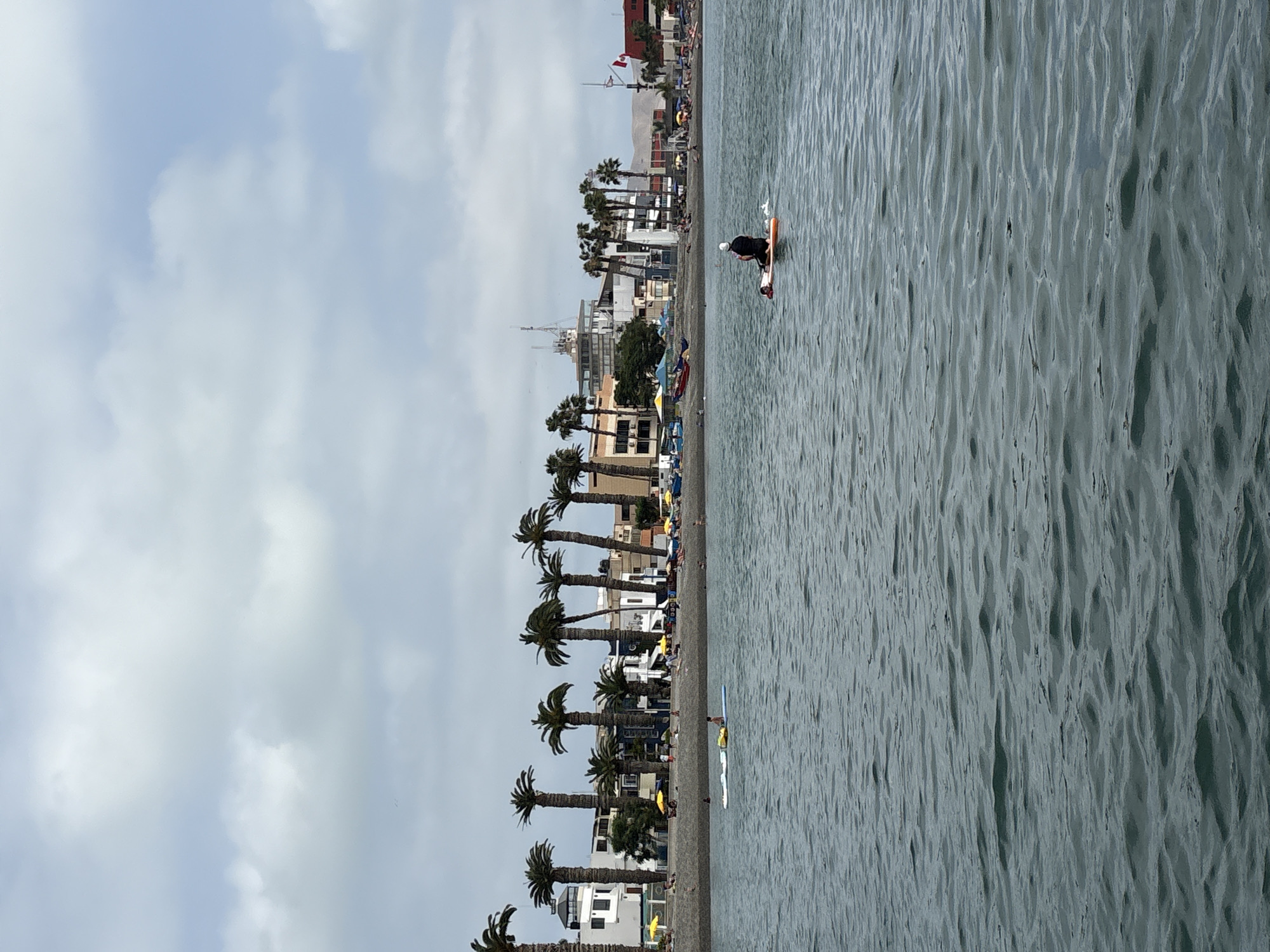 The image depicts a body of water with a shoreline in the background, characteristic of La Punta, Peru. Several palm trees line the shore, with buildings and structures situated behind them. The water is choppy, indicating a slight breeze or current. In the foreground, a person is visible on a paddleboard, facing away from the camera. Further in the distance, other individuals are present on the shore and in the water, some appearing to be on paddleboards as well. Beach umbrellas and what appear to be boats are also visible along the shoreline, suggesting a populated recreational area. The sky is overcast with scattered clouds.