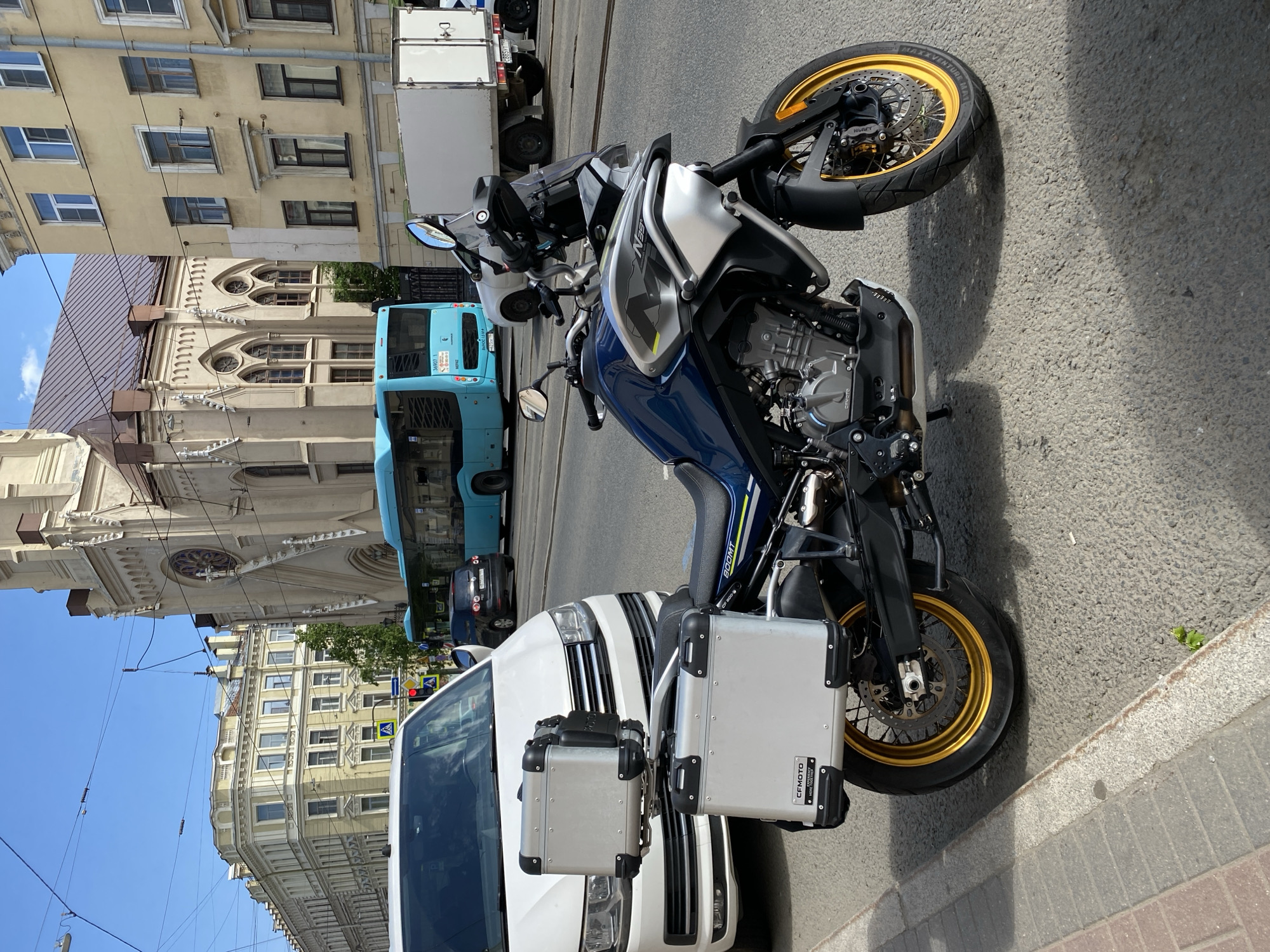 The image captures a vibrant daytime street scene in what appears to be a European city, with a prominent motorcycle in the foreground. The photo is oriented vertically, but the scene itself is horizontal.
In the immediate foreground, a blue and silver CFMOTO 800MT motorcycle is parked on the street. It features gold-colored wheel rims, black engine components, and two silver hard-shell panniers on either side, one bearing the "CFMOTO" logo. The motorcycle's tire visibly reads "MAXXVENTURE." To the left of the motorcycle, a white vehicle, possibly a large van or SUV, is partially visible.
Behind these vehicles, the street continues with what appear to be tram tracks embedded in the asphalt. A blue public transport bus is seen further down the street, displaying Cyrillic text including "М003" and "ВЫСОЦ 19 АВТ" on its rear. Other vehicles and traffic signals are discernible in the distance.
The background is dominated by historical architecture. To the left, a multi-story, light yellow building with numerous windows lines the street. In the center, a grander, lighter-colored building with intricate architectural details, including arched windows, decorative spires, and cornices, resembles a church or cathedral. Overhead wires, characteristic of electric public transport systems like trams or trolleybuses, crisscross the clear blue sky.
The scene depicts a sunny day with clear weather, suggesting mid-day or early afternoon. The overall impression is that of a bustling city environment, with a blend of modern vehicles and classic urban architecture.