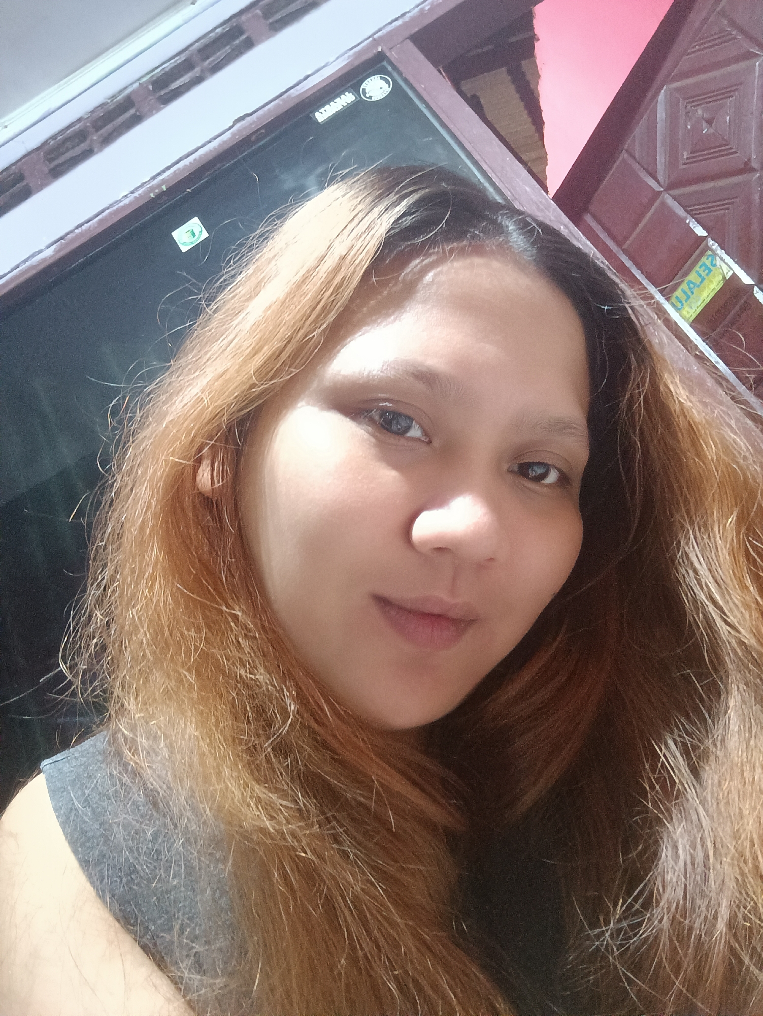 A medium shot selfie of a young woman with long, reddish-brown hair, taken indoors with a pink and brown patterned wall in the background. She is looking directly at the camera, her face slightly tilted. The image appears to have been taken in Depok, Indonesia.