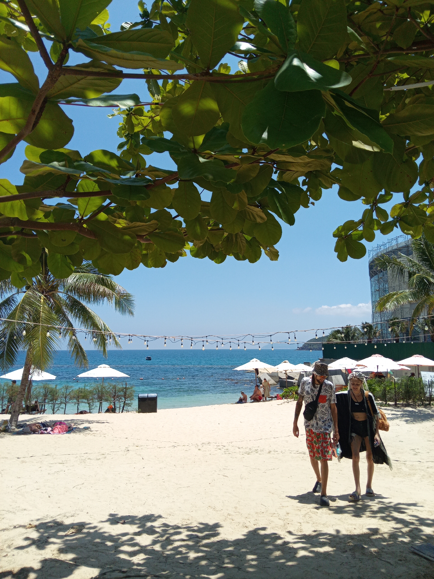 The image depicts a sunny beach scene in Nha Trang, Vietnam, looking out towards the ocean. The foreground is dominated by the dark green foliage of a tree, partially obscuring the clear blue sky. Below, a sandy beach stretches into the mid-ground.
Two individuals are walking towards the viewer on the sand in the mid-foreground. The person on the left is male, wearing a patterned bucket hat, sunglasses, a light-colored patterned short-sleeved shirt, red watermelon-print shorts, and dark slip-on shoes. He carries a dark fanny pack across his chest. The person on the right is female, wearing a patterned headscarf, a dark crop top, dark shorts, a long dark lightweight cover-up, and light-colored sandals. She carries a light brown bag over her right shoulder. Both individuals cast distinct shadows on the sand, which is also mottled with dappled shade from overhead trees.
To the left, a palm tree stands near the water's edge. Multiple white beach umbrellas are scattered along the sandy and rocky shoreline. Several other individuals are visible further back on the beach, some seated or standing near the water under umbrellas. A string of bare light bulbs hangs horizontally across the scene above the beach. The turquoise ocean is visible beyond the beach, extending to a distant horizon. A small boat is present in the water, and several buoys mark areas in the sea. In the far distance, a landmass or island is visible on the left. On the right, a large multi-story building under construction, partially covered in scaffolding, dominates the skyline, suggesting a developing resort area.