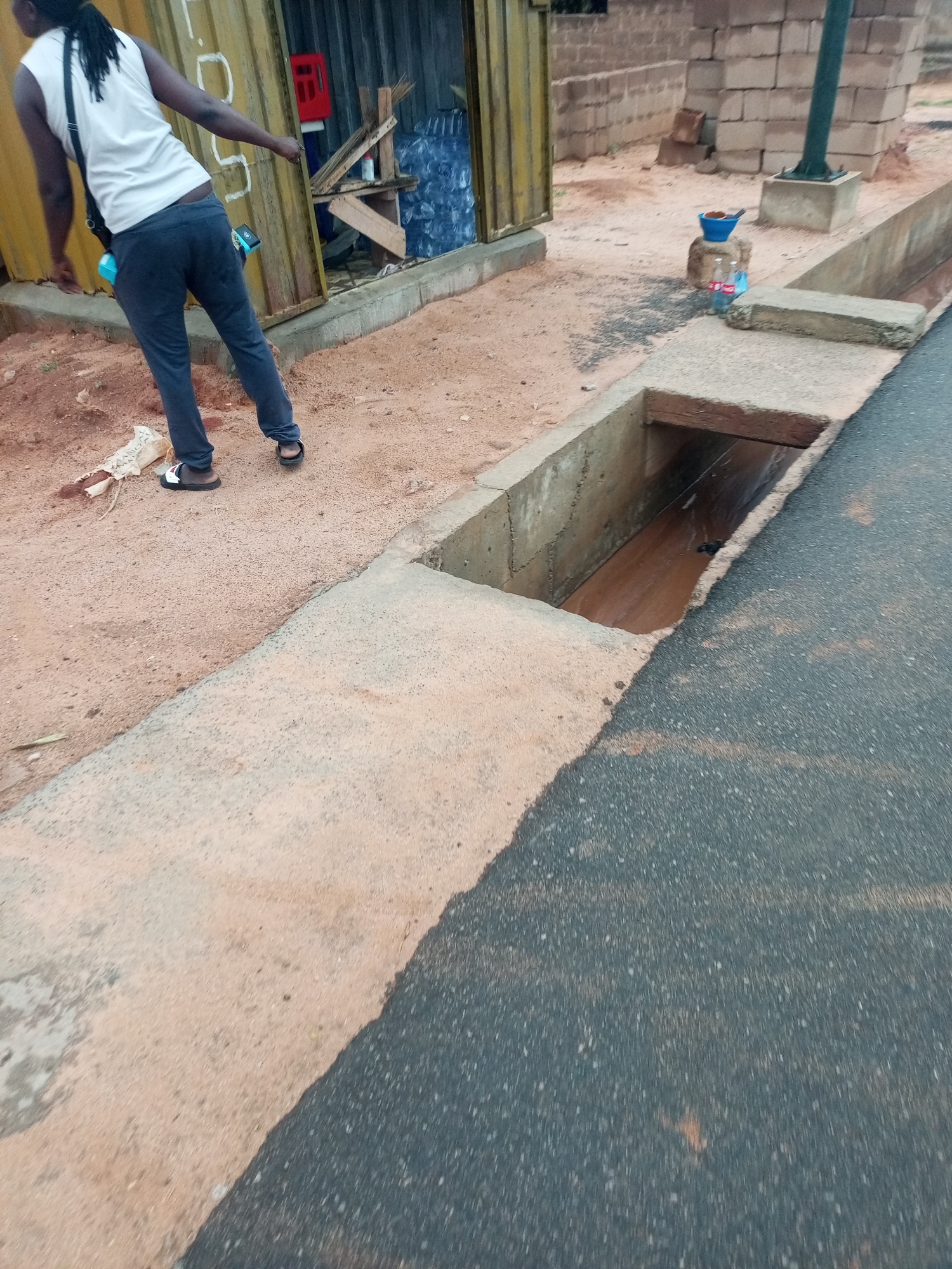 A person in a white shirt and dark pants is standing in front of a yellow building in Gombe, Nigeria. The person is near an open drainage ditch running alongside a paved road. A blue bucket is positioned on a rock near some bottled drinks next to the building.
