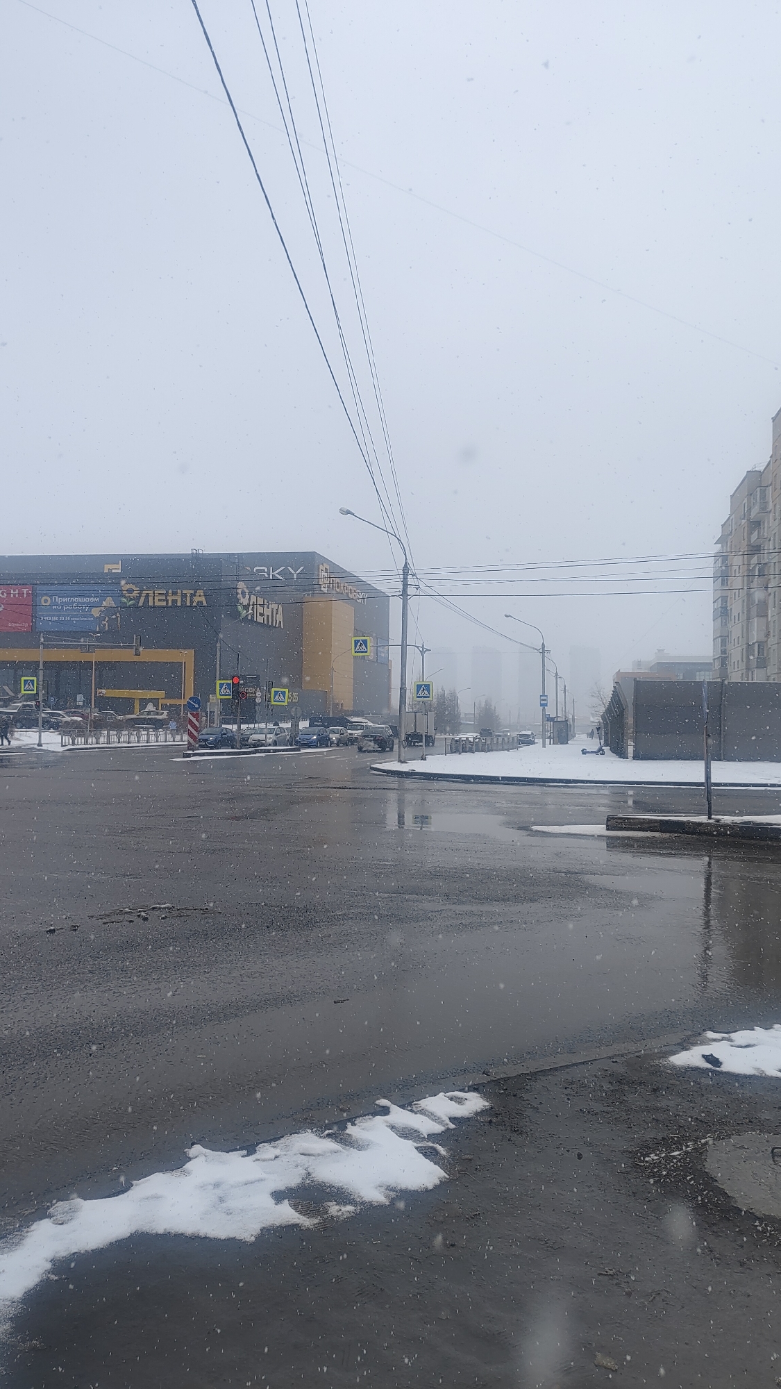 The image depicts a city intersection in Krasnoyarsk, Russia, on a snowy, overcast day. Light snowflakes are visible throughout the frame, falling onto the wet, dark asphalt roads, which show reflections of the dull sky. Patches of white snow line the curbs and are scattered on the ground, indicating recent or ongoing snowfall.
On the left side of the intersection, a large commercial building stands prominently, identified by bright yellow Cyrillic lettering against a dark facade. The main sign reads "ЛЕНТА" (LENTA), a Russian hypermarket chain, with "SKY" also visible above it. Another sign below "ЛЕНТА" appears to say "ПРИГЛАШАЕМ НА РАБОТУ" (We invite you to work/hire).
At the intersection in front of the LENTA building, cars are present, some parked, others on the road. A red traffic light is illuminated, and several yellow, A-frame pedestrian crossing signs are placed strategically. Overhead, numerous power lines stretch across the sky.
To the right, a road extends into the hazy distance, where faint outlines of tall buildings are barely visible through the snow and fog. Along this road, a street lamp and a bus stop sign are distinguishable. On the far right, a multi-story residential building with many balconies completes the urban scene.
The time of day appears to be daytime, but the heavy cloud cover and snow create a flat, muted light, contributing to a cold and damp winter atmosphere. No specific human activity beyond traffic movement is clearly highlighted, though a pedestrian is vaguely visible on the far left.