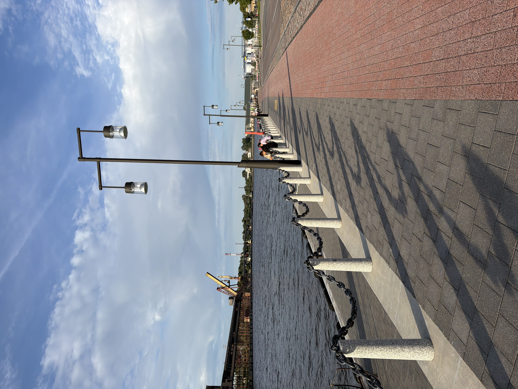 The image depicts a modern waterfront promenade in Ventspils, Latvia. The promenade is paved with dark gray rectangular bricks and a section of red brick paving. It is lined with granite bollards and a heavy chain railing separating it from the dark, rippling water of a harbor or river. Beyond the railing, the water stretches towards an industrial area featuring large buildings and cranes, including a prominent yellow construction crane.
Several streetlights with cylindrical glass shades are evenly spaced along the promenade. In the mid-ground, a small group of people, including children, are visible walking and interacting near the railing, casting long shadows on the pavement. Further down the promenade, more streetlights and distant figures can be seen. The sky is partly cloudy with white clouds against a blue background. The overall scene suggests a well-maintained public space designed for recreation and viewing the harbor.