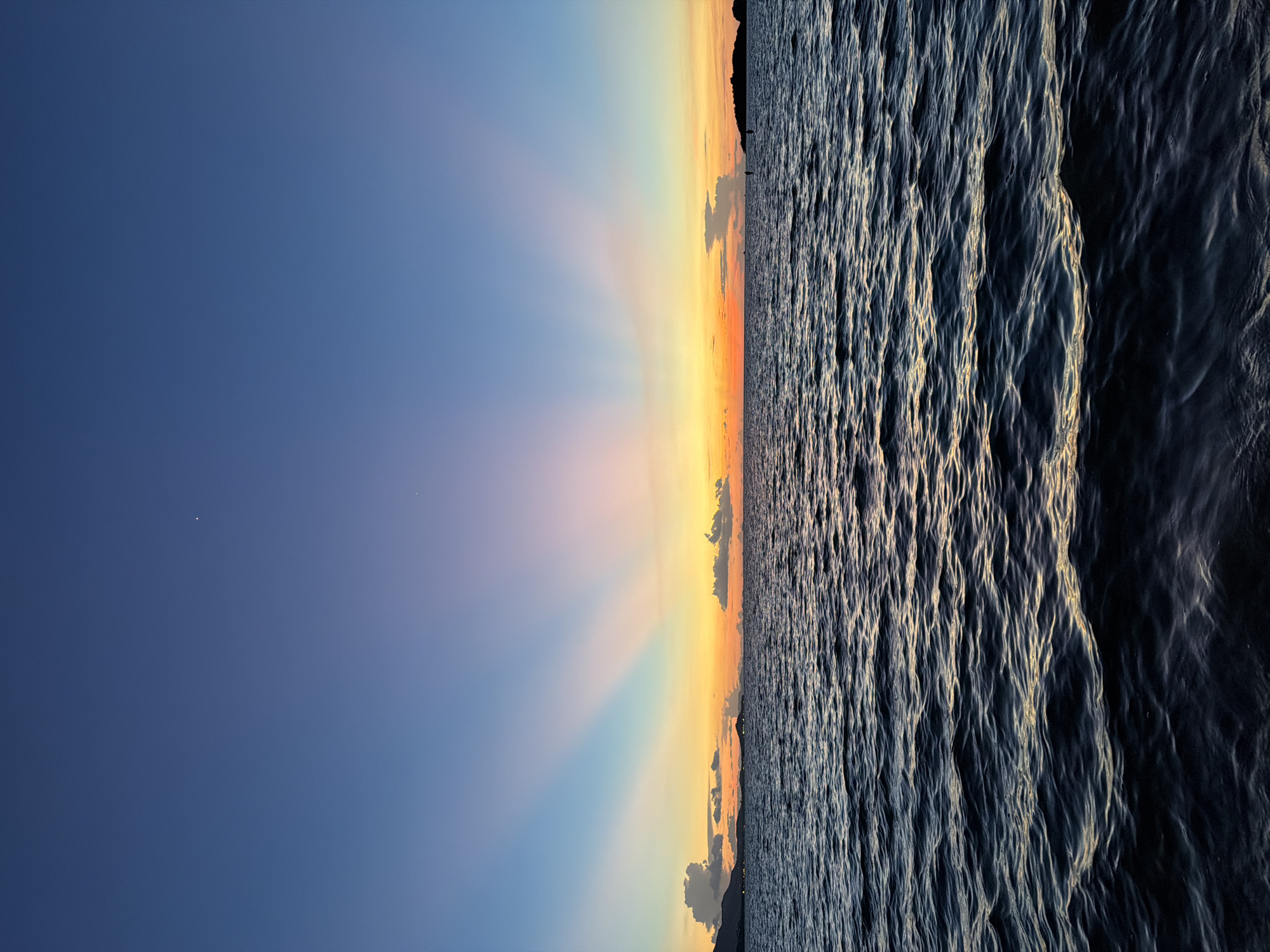 This is a vertical, wide-angle photograph of a sunset over the ocean at Baan Plai Laem, Thailand. The image is divided into three horizontal bands: the sky, the horizon with land, and the ocean. The sky is a gradient of deep blue at the top, transitioning to pale blue, then to yellow and orange near the horizon. Rays of light fan out from the horizon, creating a dramatic visual effect. A few clouds are scattered in the sky, particularly near the horizon.
The horizon is marked by a dark silhouette of a hilly coastline, with what appears to be a few distant lights from a settlement. The ocean occupies the bottom portion of the image and is characterized by choppy waves. The setting sun casts a golden light on the crests of the waves, creating a stark contrast with the darker troughs. The overall mood is serene and tranquil, capturing the beauty of a tropical sunset. The time of day is clearly evening, as indicated by the sunset and the soft, warm lighting. There is no visible text in the image.