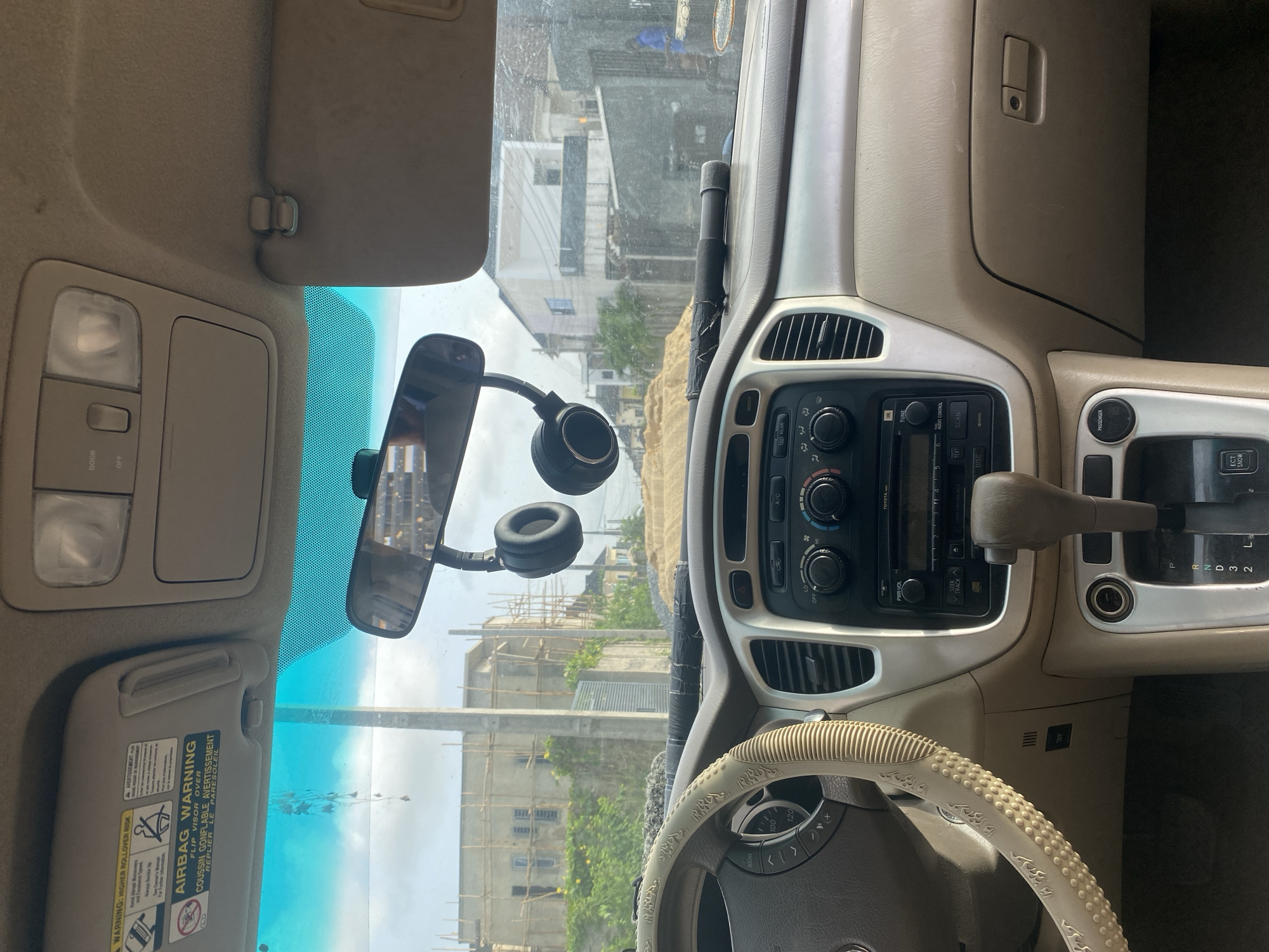 The image shows the interior of a Toyota SUV. A pair of headphones are hanging from the rearview mirror. The car is parked facing a street with buildings under construction.