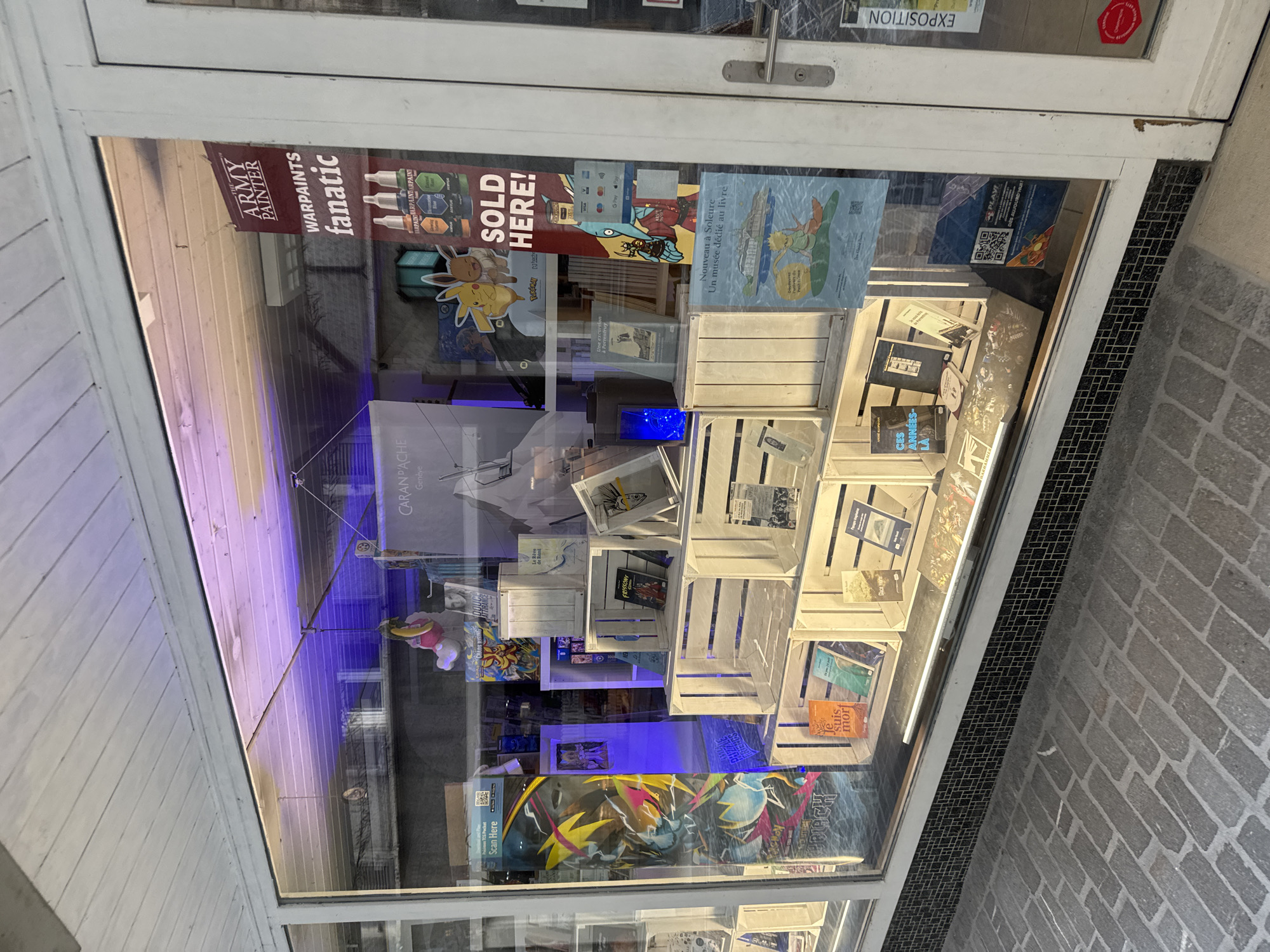 The image shows a storefront window display for a bookstore in Porrentruy, Switzerland. The display features a variety of books, including art books, novels, and graphic novels, arranged on white wooden crates. There are also posters and other promotional materials visible. The lighting in the store is purple and blue. The exterior of the store has a brick sidewalk. The store appears to be closed, as no customers are visible. A sign that says "CARAN D'ACHE Genève" is visible in the window. There is also a sign that says "THE ARMY PAINTER warpaints fanatic". A sign also says "SOLD HERE!". There are also signs for "VISA", "MasterCard", and "Apple Pay". Another sign says "Nouveau à Soleure - Un musée dédié au livre". A poster on the right side says "EXPOSITION".
