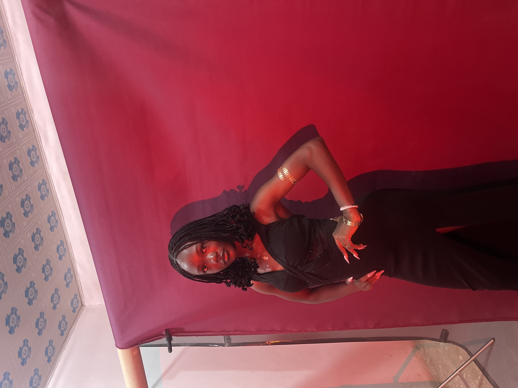 A young Black woman with braided hair stands against a red backdrop in what appears to be an indoor studio setting in Gura Top, Nigeria. She is wearing a long, sleeveless black dress with a thigh-high slit. The dress has a v-neckline and intricate beading or detailing around the bodice. She is adorned with gold jewelry, including an arm cuff on her left arm and bracelets and rings on her right hand. Her nails are long and painted in a bright pink color. She is posing with her hands on her hips, and her body is angled slightly towards the camera. A shadow of her figure is cast on the red backdrop behind her. The ceiling above is visible and features a white surface with a repetitive blue and white patterned design. To the left of the woman, a lighting stand is partially visible, suggesting a photographic setup. The lighting in the room casts a red hue on the scene, particularly on the woman and the backdrop.