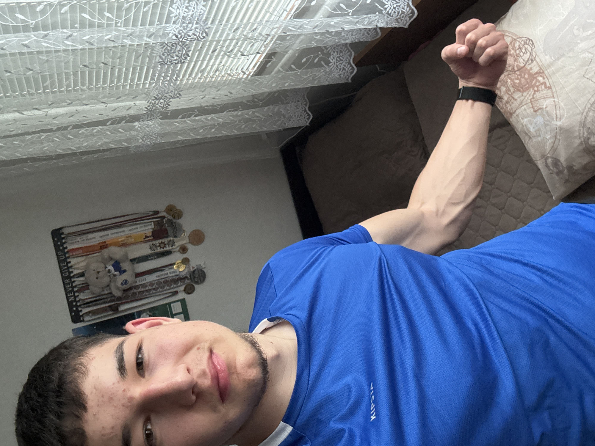 A young man is taking a selfie indoors. He is wearing a blue Kipsta brand athletic shirt. He is flexing his right arm, showing prominent veins. He is wearing a black wristband. Behind him is a bed with a patterned pillow and a plain brown comforter. To the left, on the wall, are medals and a teddy bear hanging. A window with lace curtains is visible in the background. The photo was likely taken in a residential setting, but there is nothing that confirms the city of Zlatograd, Bulgaria.