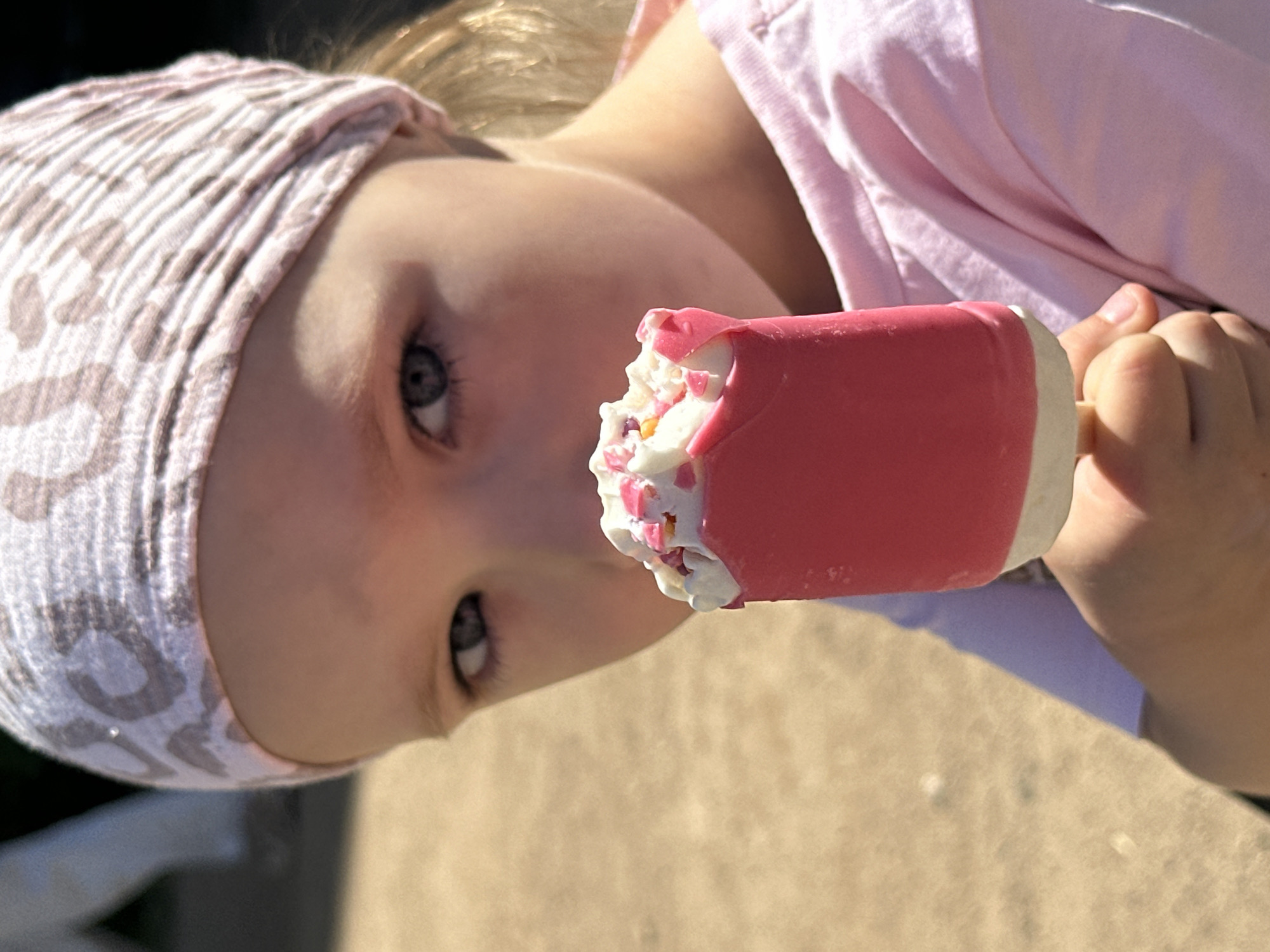 A young child with light skin, blue-grey eyes, and a white and pink patterned headband is eating a pink popsicle with white frosting and colorful sprinkles. The child is wearing a light pink shirt. The background is blurry and appears to be an outdoor setting, possibly a sidewalk. The image appears to be taken in the city of Chaikovsky, Russia, based on external information not provided in the image.