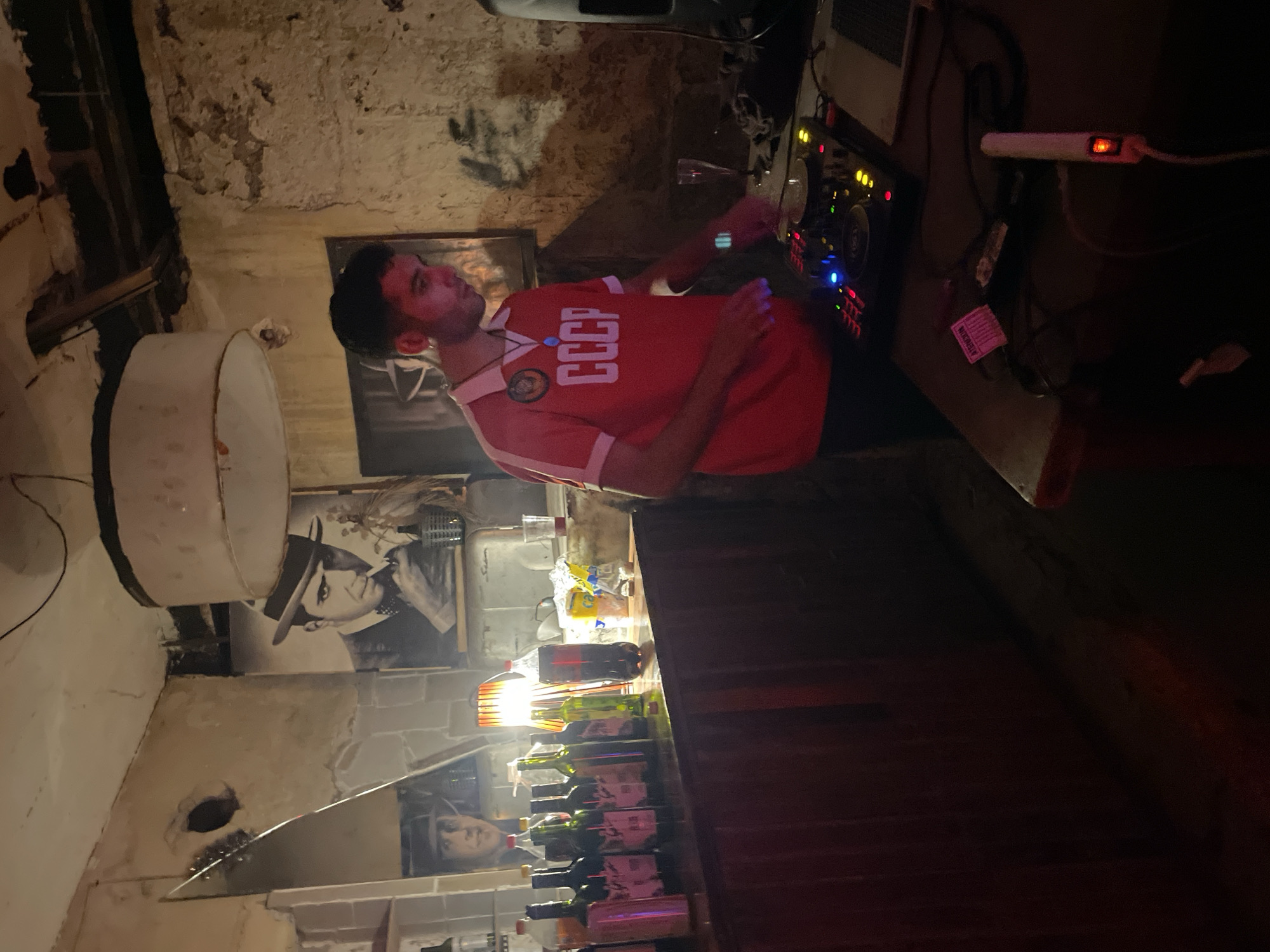 A man stands inside a building or bar, likely in Buenos Aires, Argentina, based on the architectural style and cultural references. The building appears to be old, with weathered walls and exposed brick. The man is wearing a red shirt with "CCCP" written on it. He is positioned behind a bar that is lined with bottles. Beside him are turntables and an amplifier. There are images on the wall behind the bottles.