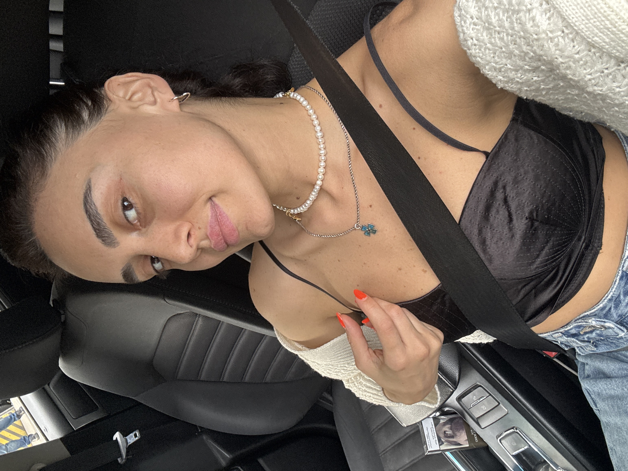 A woman is sitting in the driver's seat of a car, with her body angled towards the camera. She has dark hair pulled back and is wearing a black satin bra, a pearl necklace, and a white knitted cardigan draped over her shoulders. Her fingernails are painted bright orange. A black seatbelt crosses her chest. The interior of the car is visible, including the dark leather seats and the center console with car controls. The woman is looking directly at the camera with a slight smile. The scene appears to be outdoors, possibly during the day, as indicated by the lighting. The location context is Tbilisi, Georgia. There is no visible weather information. A pack of cigarettes with Russian text is visible on the center console.
