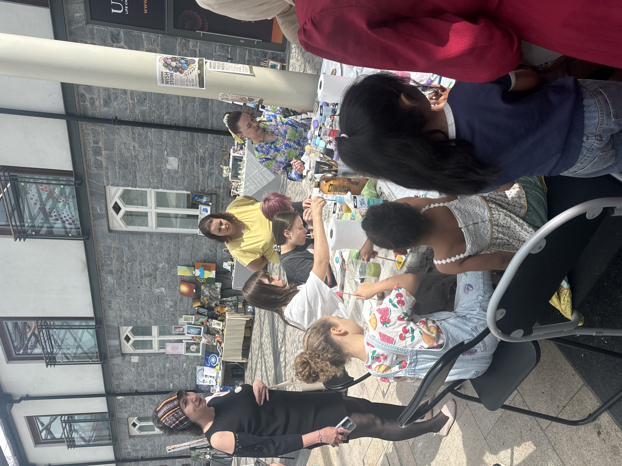 In Kilkenny, Ireland, a group of people, including adults and children, are gathered around a table outdoors, engaged in what appears to be an art or craft activity. The setting is paved, suggesting a public space like a street or market. Behind the group, a building with stone facade and multi-story windows is visible, characteristic of Kilkenny's historic architecture. A white pillar stands to the right of the frame. Several tables with various items, including paintings, art supplies, and decorations, are set up in the background, indicating a potential market or festival. The people are focused on their tasks, with some holding brushes and small objects, possibly painting stones or other craft items. The overall atmosphere seems relaxed and creative.