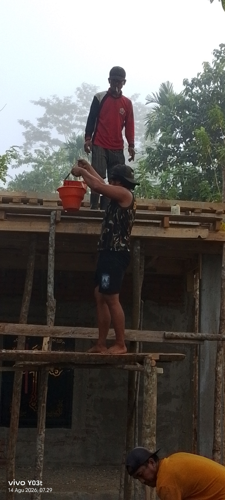 The image captures a scene of construction work in Woiha, Indonesia. Two men are visible on a wooden scaffolding structure under a foggy sky. One man, standing higher on the scaffolding, wears a red and black long-sleeved shirt and striped pants, with a cigarette in his mouth. He appears to be supervising. Below him, another man, wearing a patterned sleeveless shirt, black shorts, and a dark cap, is holding an orange bucket in his hands, seemingly in the process of transferring something or preparing to pass it up. He is barefoot and standing on the wooden planks. The construction site is characterized by unfinished wooden beams and support structures. In the background, lush green trees and vegetation are visible through the fog, indicating a natural, possibly rural or suburban, environment. The lighting suggests it is early morning or late afternoon due to the fog.