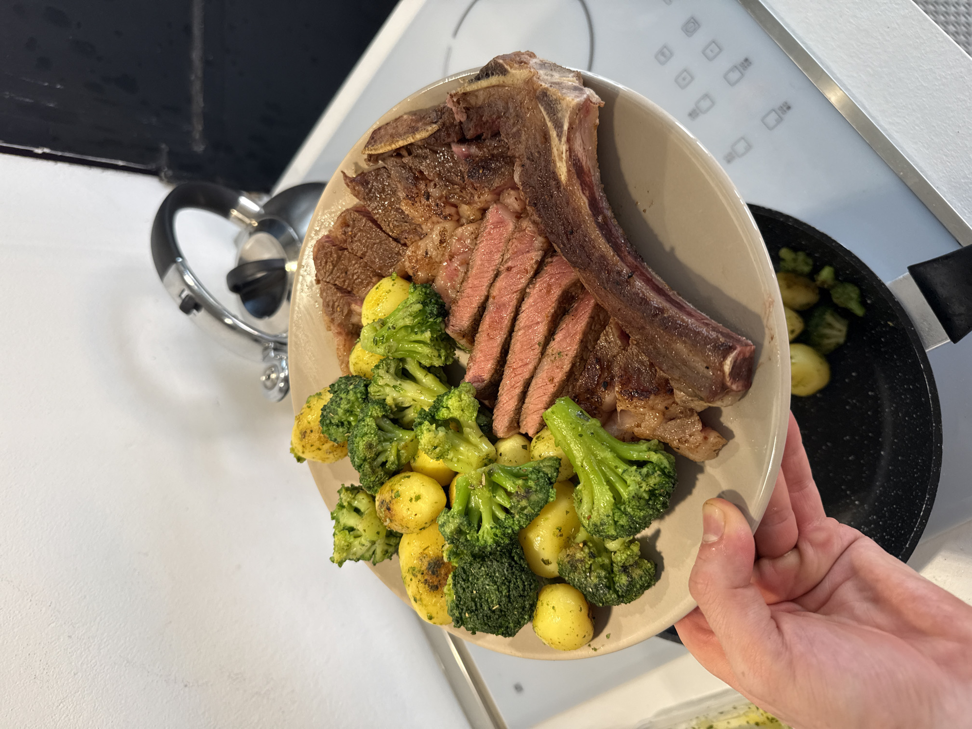 A person holds a plate of food containing a large, sliced T-bone steak, broccoli florets, and small yellow potatoes. The steak has a significant bone attached, and the slices reveal a medium-rare interior. Some of the vegetables appear to be seasoned with herbs. In the background, a stainless steel kettle sits near a black-and-white wall. A frying pan containing similar broccoli and potato mix is also visible on a white induction hob. The scene suggests a kitchen environment. There is no evidence to confirm or deny the location is Milan, Italy.
