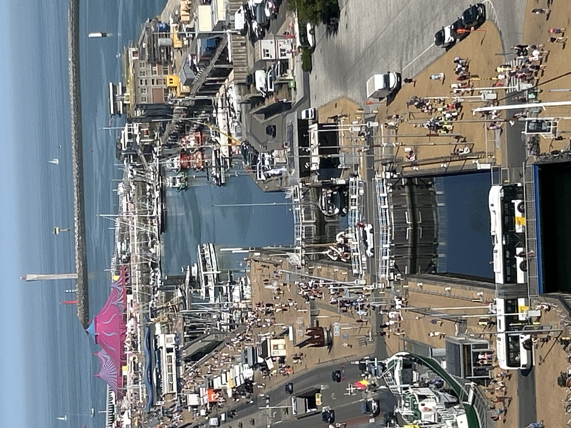 This is a high-angle, wide shot of the port of Ostend, Belgium, on a clear, sunny day. In the foreground, a large, blue-water lock system separates the inner harbor from the outer sea. The lock is filled with water, reflecting the sky.
On the left side of the lock, a promenade is lined with people strolling and congregated. Several boats, including a large green and white vessel, are docked along the promenade. The promenade itself is paved with yellow bricks.
To the right of the lock, a paved area features several parked cars and a white van. A small, white building with a red roof stands near the parked vehicles. Further to the right, a paved street is visible, with more cars and a black taxi.
In the background, a vibrant pink and purple circus tent stands out, suggesting an event or attraction. Beyond the tent, a harbor filled with numerous sailboats and other vessels stretches towards the horizon. A breakwater separates the harbor from the open sea, where a lighthouse and a red lighthouse are visible. The sea is a deep blue, meeting a pale blue sky. The overall scene depicts a bustling port area with a mix of commercial activity, leisure, and maritime elements.