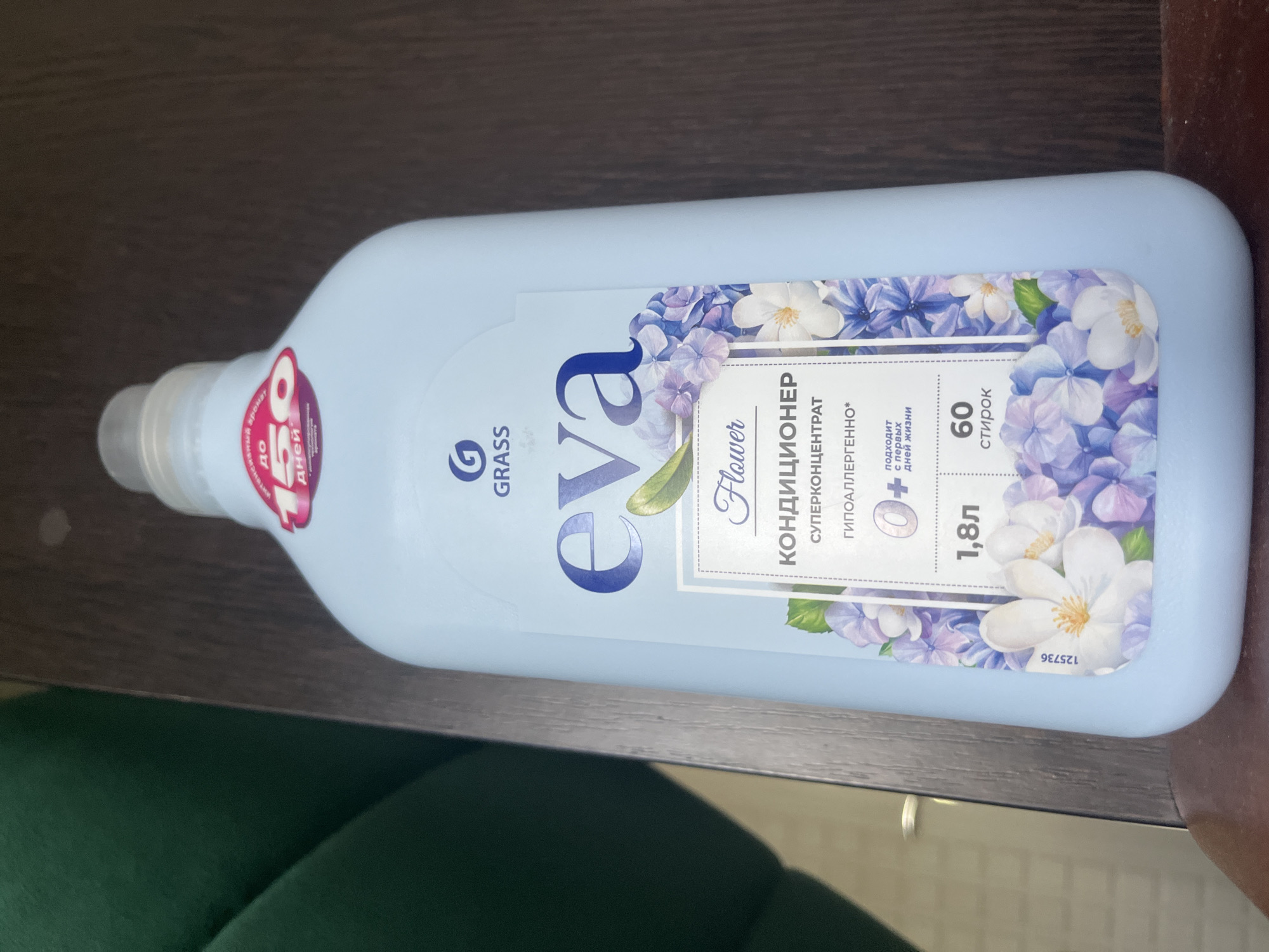 The image shows a light blue plastic bottle of "eva Flower" fabric conditioner made by GRASS. The bottle is light blue and has floral imagery, primarily hydrangeas and white flowers, on its label. Text on the label indicates it is a super-concentrated conditioner, hypoallergenic, and suitable from the first days of life. It contains 1.8 liters and provides 60 washes. A sticker on the bottle reads "до 50 дней" (up to 50 days) related to fragrance duration. The bottle is placed on a dark wooden surface. In the background, a portion of a dark green upholstered object is visible. The scene is an indoor setting, but specific location details within Volzhsky, Russia, are not discernible from the image alone.
