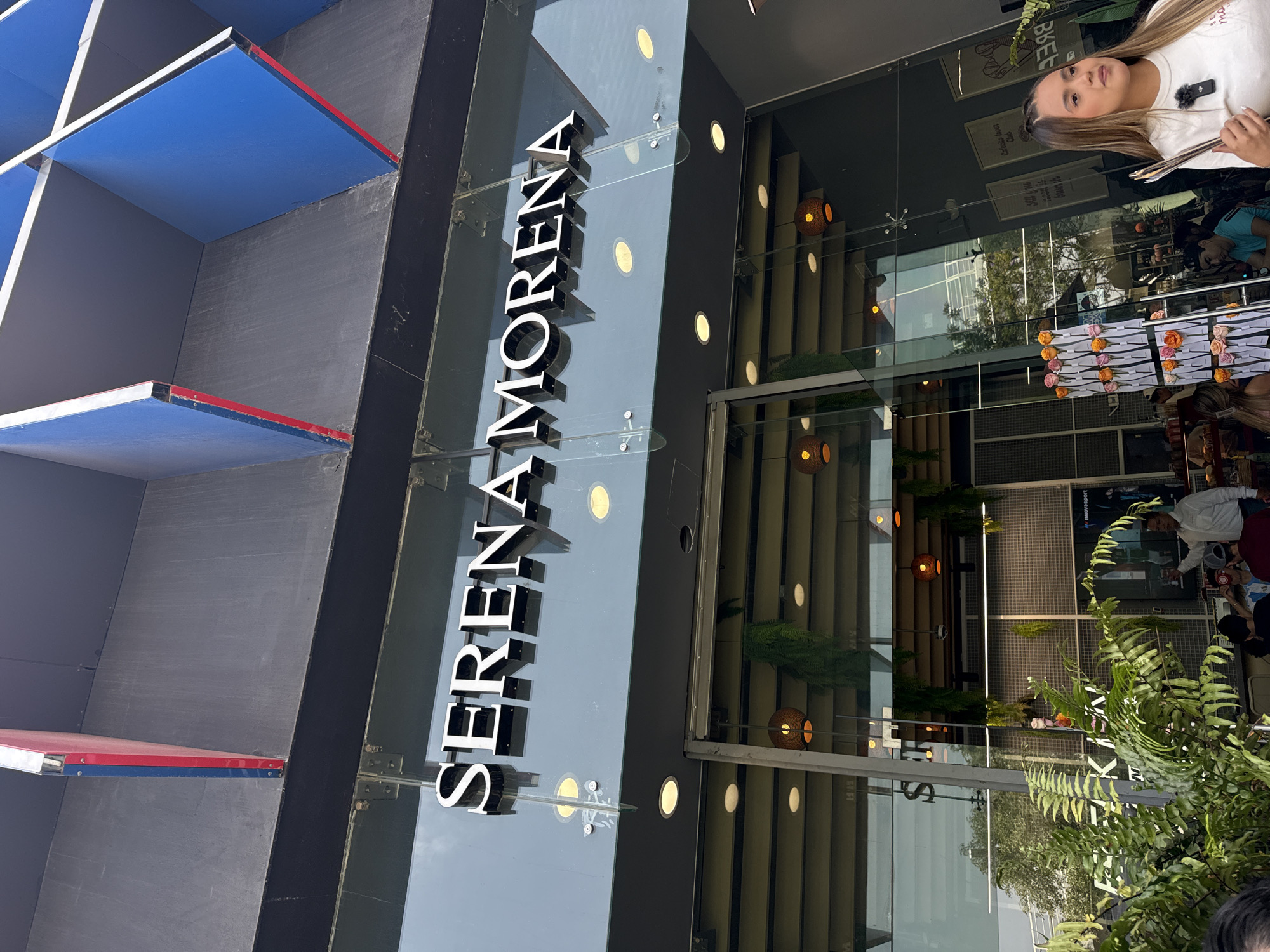Here's a description of the image:
**Scene Overview:**
The image shows the exterior of a restaurant named "SERENA MORENA" in Puebla, Mexico. The restaurant has a modern facade with a geometric, architectural design.
**Architectural Details:**
The building features a mix of materials including glass, metal, and painted sections. The structural design incorporates a series of recessed, shelf-like elements painted in colors, including blue and gray, with red accents. The restaurant’s name is displayed vertically in large, white block letters attached to the building's exterior.
**People and Activity:**
There are people both inside and outside the restaurant. A woman is seen at the edge of the frame, partially reflected in the glass windows of the restaurant, as well as an indistinct view of patrons seated at tables within the dining area, partially obscured by foliage.
**Environment:**
The lighting suggests it's daytime. The overall impression is of a modern, upscale restaurant in an urban environment. There is foliage and greenery around the entrance, giving the restaurant a touch of nature.
**Location:**
Puebla, Mexico.
**Key Elements:**
* "SERENA MORENA" sign
* Modern building facade
* Geometric architecture
* People inside and outside the restaurant
* Foliage