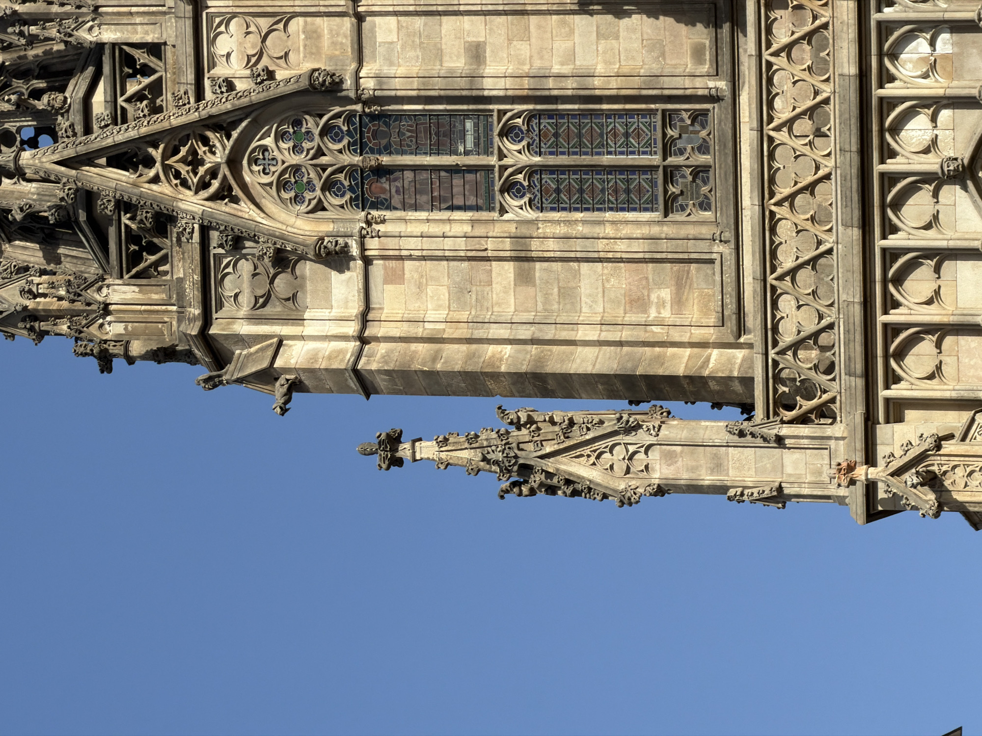 This is a close-up, low-angle outdoor, vertical shot of the exterior architecture of a Gothic-style building. The building is constructed of light-colored stone blocks and features intricate carvings and architectural details. In the foreground and to the right, a large section of a tower is visible, adorned with pointed arches, decorative tracery, and stained glass windows. Two pointed windows with multi-colored stained glass are prominent. Above these windows, more elaborate Gothic ornamentation can be seen, including small gargoyle-like figures projecting from the stonework. To the left, another smaller, pointed spire also covered in detailed carvings rises against a clear, bright blue sky. The sunlight casts shadows, highlighting the textured surfaces of the stone. The overall impression is one of ancient, detailed craftsmanship. This is the Cathedral of Barcelona, located in Barcelona, Spain.