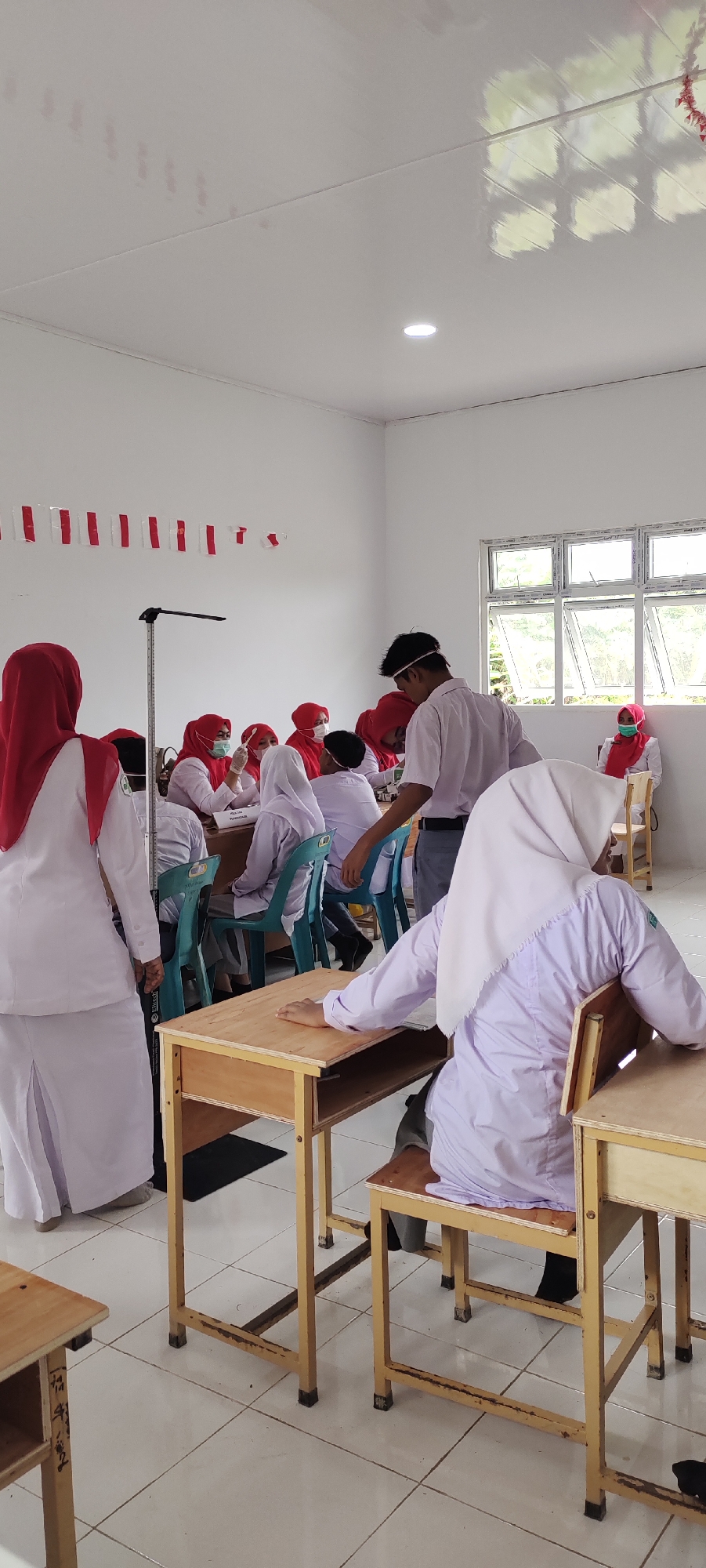 In a classroom in Langnibong, Indonesia, students wearing white uniforms and red hijabs are seated at wooden desks. A male student wearing a white uniform and a face mask stands and addresses the class. The walls of the classroom are white and decorated with red and white Indonesian flags. Large windows allow natural light to enter the room, and the floor is tiled. The scene appears to depict a school event or lesson.