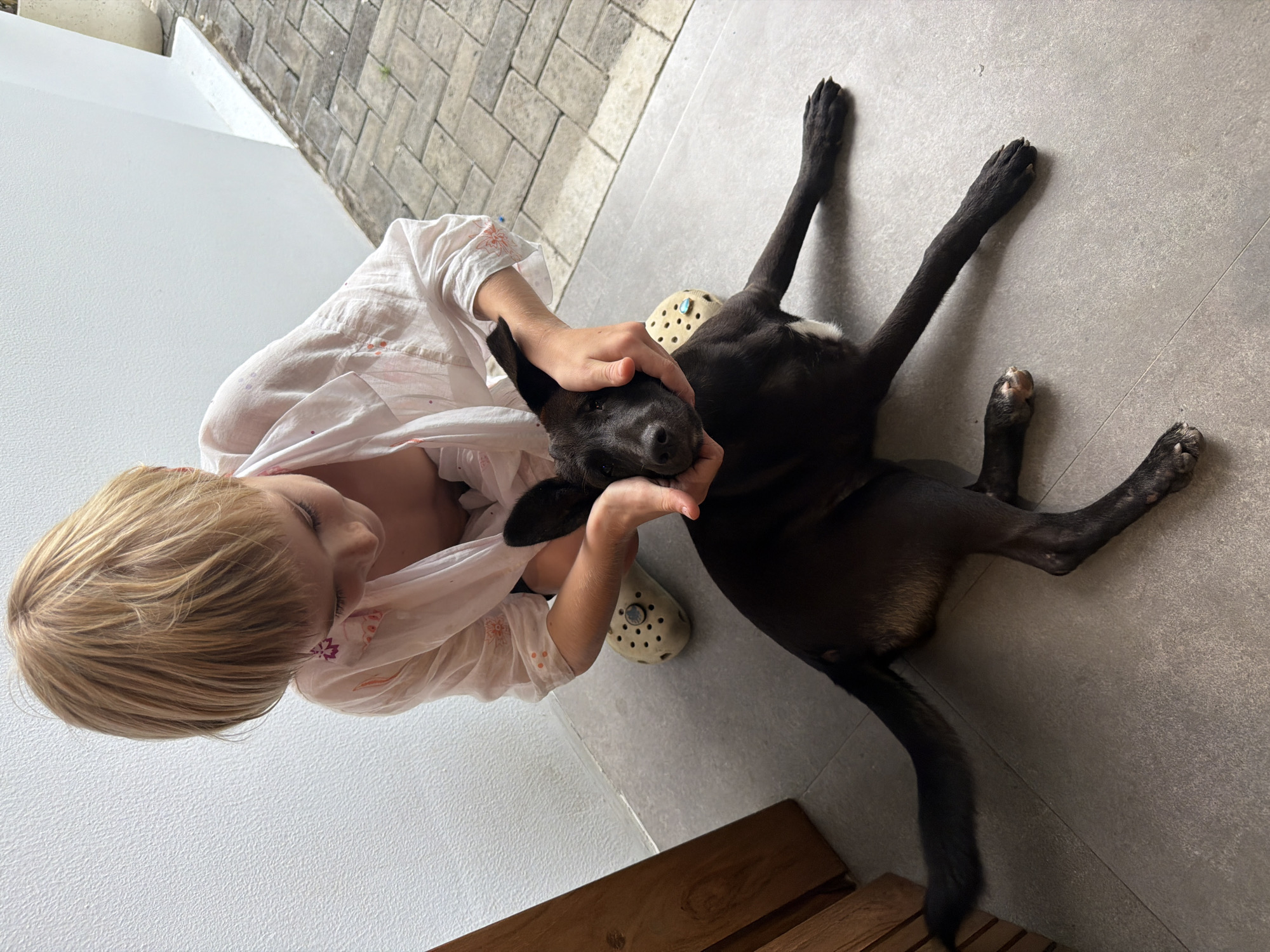 A child with blonde hair is crouching and interacting with a black dog. The child is wearing a light-colored, short-sleeved shirt. The dog is lying on its back on a tiled floor. The child is holding the dog's head in their hands. The dog is lying near a pair of shoes and a wooden structure. The photo appears to be taken outdoors, near a wall and a paved area. It's located in Kuta Selatan, Indonesia.