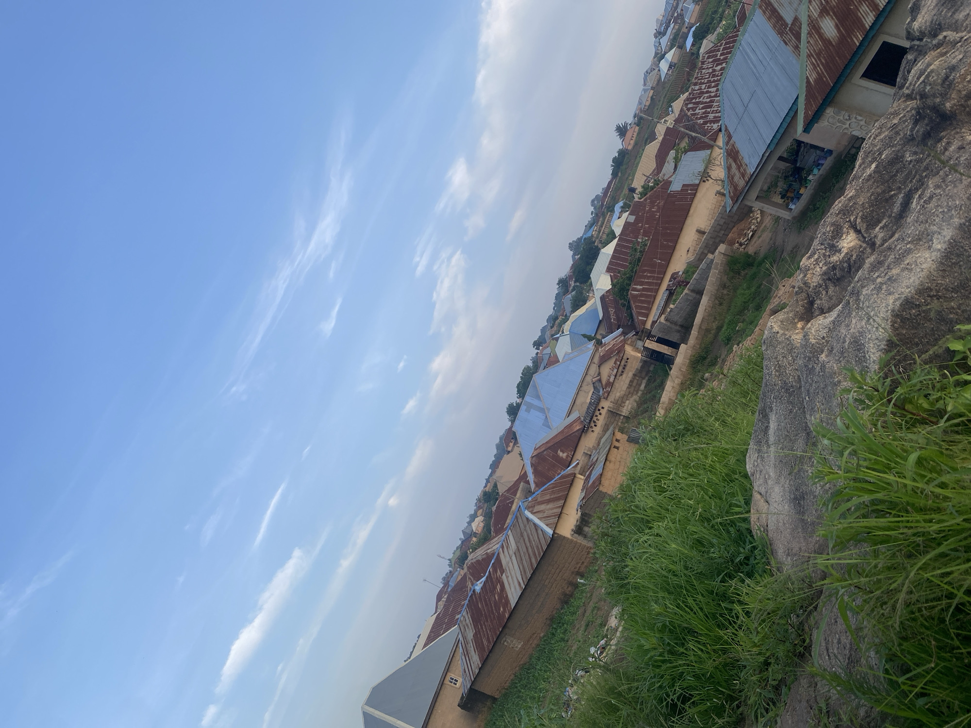 The image shows a high-angle view of a residential area in Bukuru, Nigeria. The scene is dominated by numerous houses with corrugated metal roofs, varying in color from rusted brown to light blue. The houses are built close together and are nestled on a gentle slope. In the foreground, large rocks and lush green grass are visible, suggesting a natural, slightly overgrown terrain. The sky is a clear blue with thin, wispy clouds, indicating fair weather. The overall impression is of a densely populated urban or semi-urban environment with a rustic feel, possibly a neighborhood on the outskirts of the city, given the presence of natural elements like rocks and grass. There are no people or visible signs of activity in the image.