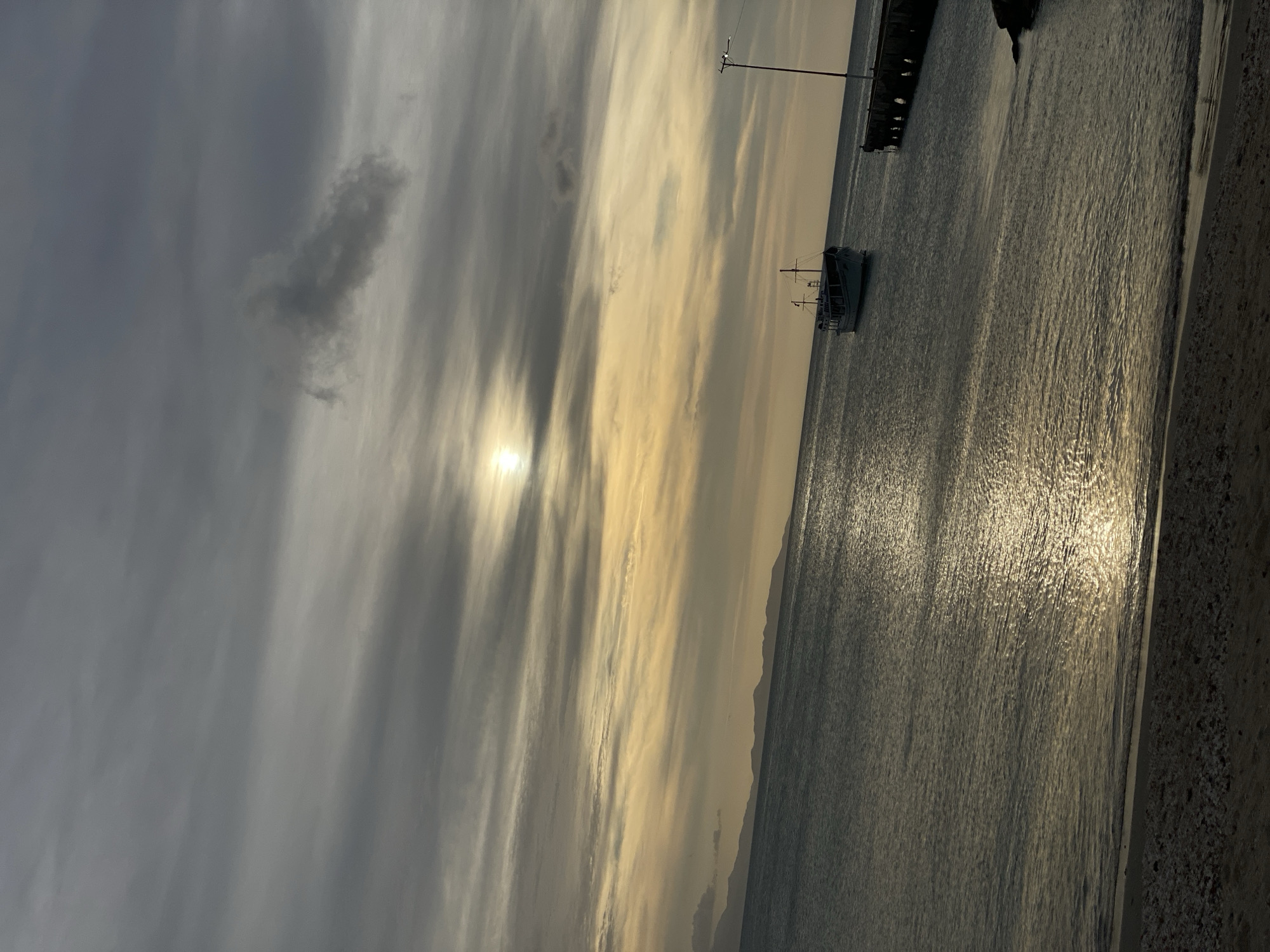 The image depicts a coastal scene in Juan Griego, Venezuela, at either sunrise or sunset. The sky is largely covered by stratocumulus clouds, presenting a gradient from dark grey in the upper portion to yellowish-orange hues near the horizon, where the sun is visible as a bright, partially obscured disc in the upper-left quadrant. A distant landmass, appearing as dark silhouettes of hills or mountains, stretches along the left half of the horizon. The calm, dark blue-grey sea occupies the middle ground, reflecting a shimmering path of sunlight from the sun towards the foreground. A white and blue, two-masted boat, featuring a visible cabin, is moored in the water in the mid-right. To its right, a wooden pier or jetty with multiple pilings extends into the water from the right edge of the frame. A single, tall light pole stands vertically on the pier. The immediate foreground shows a narrow strip of sandy beach mixed with small, light-colored pebbles or shells at the water's edge. No individuals or specific actions are discernible.