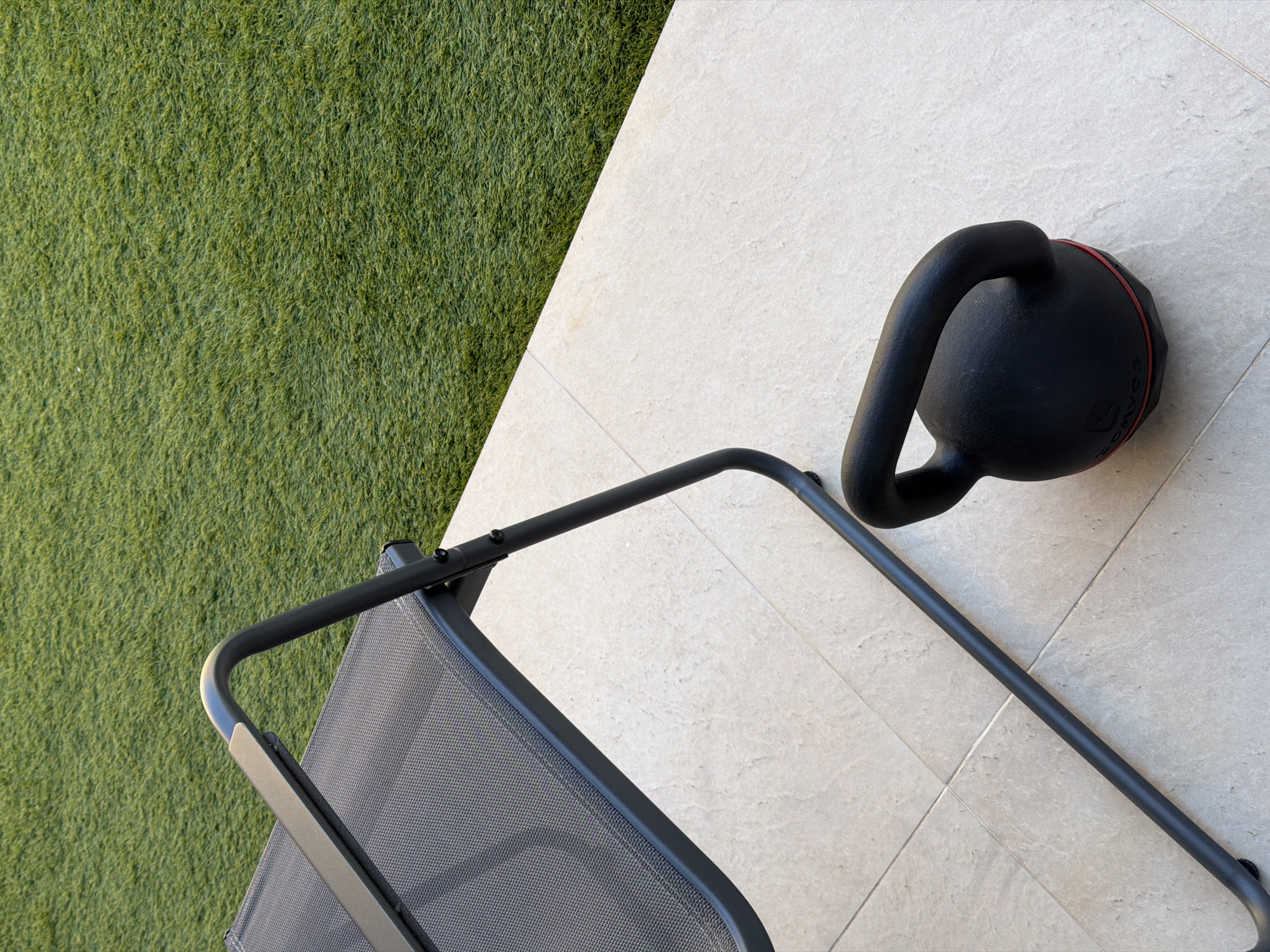 A kettlebell rests on a tiled surface next to a dark metal lounge chair. Behind the chair and to the right of the image, artificial turf covers the ground. The scene is outdoors, likely on a patio or balcony in Finestrat, Spain, as indicated by the context. No people are visible in the image, and there are no apparent actions or interactions taking place. The lighting suggests it is daytime.