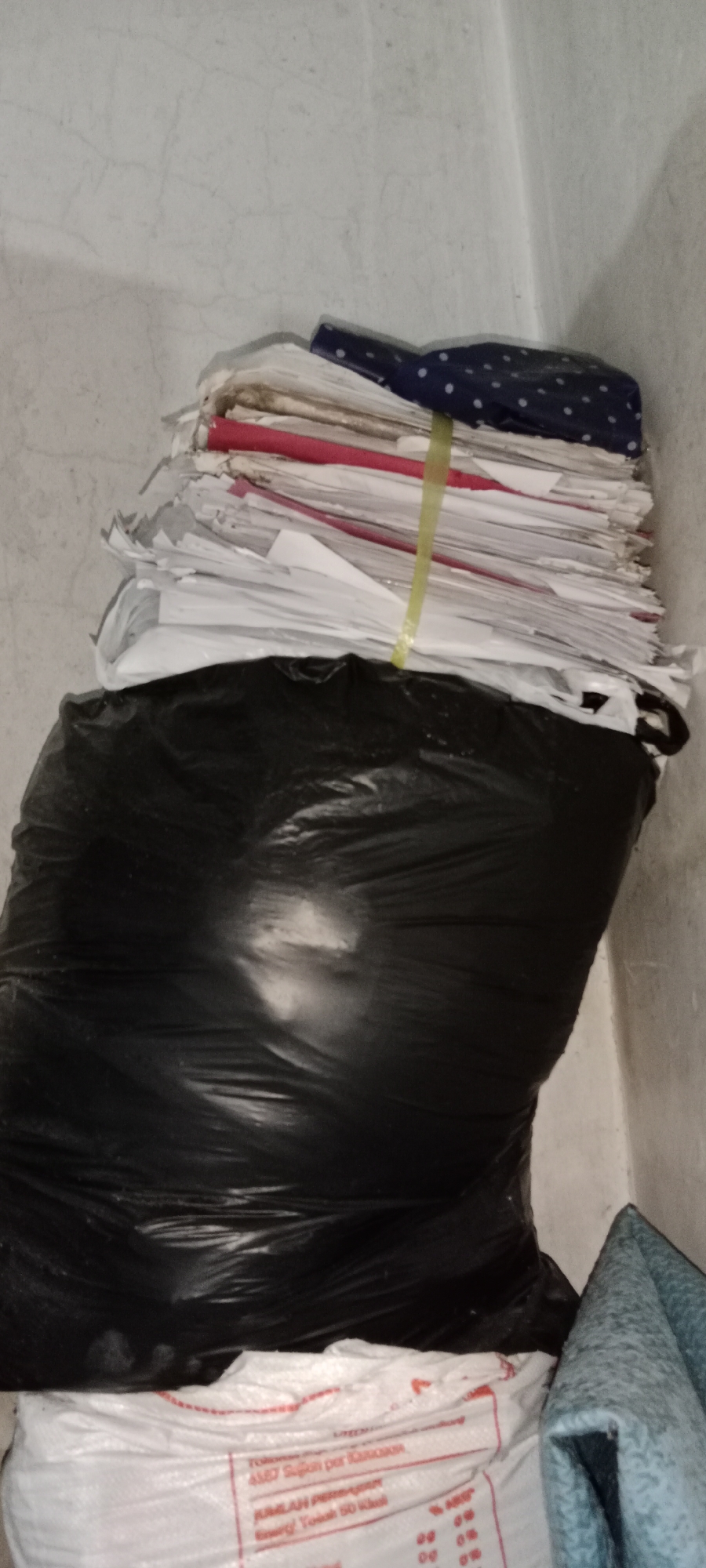 The image depicts a black trash bag filled with what appears to be crumpled papers and documents. A stack of papers is visible on top of the trash bag, secured by a yellow tie and topped with a dark blue fabric with white polka dots. Below the trash bag, a white bag with red lettering is partially visible. To the right of the image, a blue patterned fabric is draped. The background consists of a textured, off-white wall with visible cracks, suggesting an indoor environment. The lighting is dim, casting shadows and highlighting the texture of the wall and the plastic bag. The overall scene suggests a storage area or a discarded pile of materials. There is no visible indication of the specific location within Subang, Indonesia.