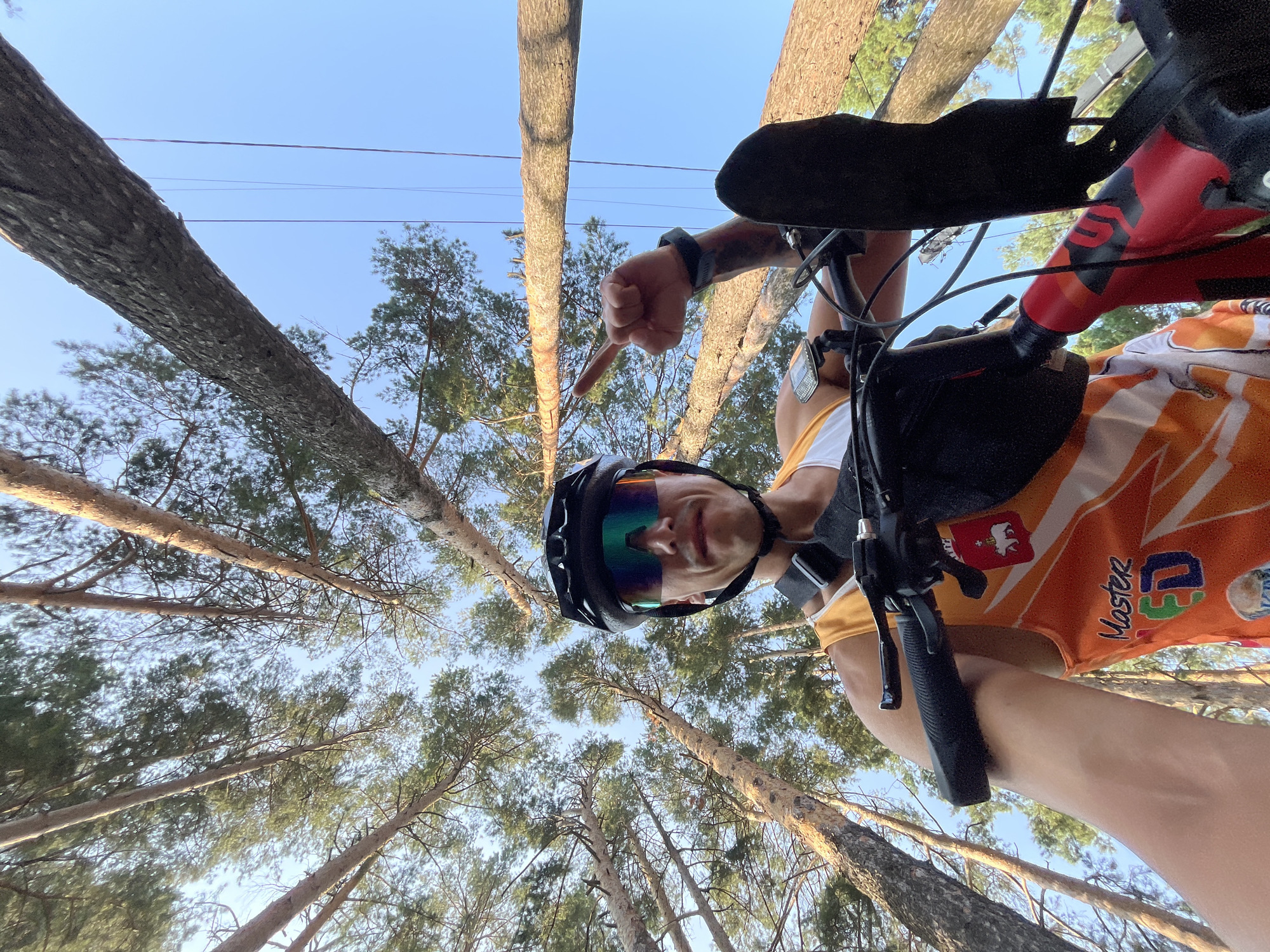 A low-angle, wide shot shows a person on a bicycle, viewed from below, looking up towards the sky. The person is wearing a black cycling helmet and iridescent sunglasses, with a white tank top underneath an orange and white sleeveless jersey featuring graphics. The jersey has a crest with a bear and shield on the left chest. The person's left arm is extended, with a finger pointing upwards towards the sky. The background is dominated by tall pine trees, with their trunks and branches extending towards a clear blue sky. The scene appears to be an outdoor environment, possibly a park or forest trail, with sunlight filtering through the trees. The location is identified as Chaikovsky, Russia.