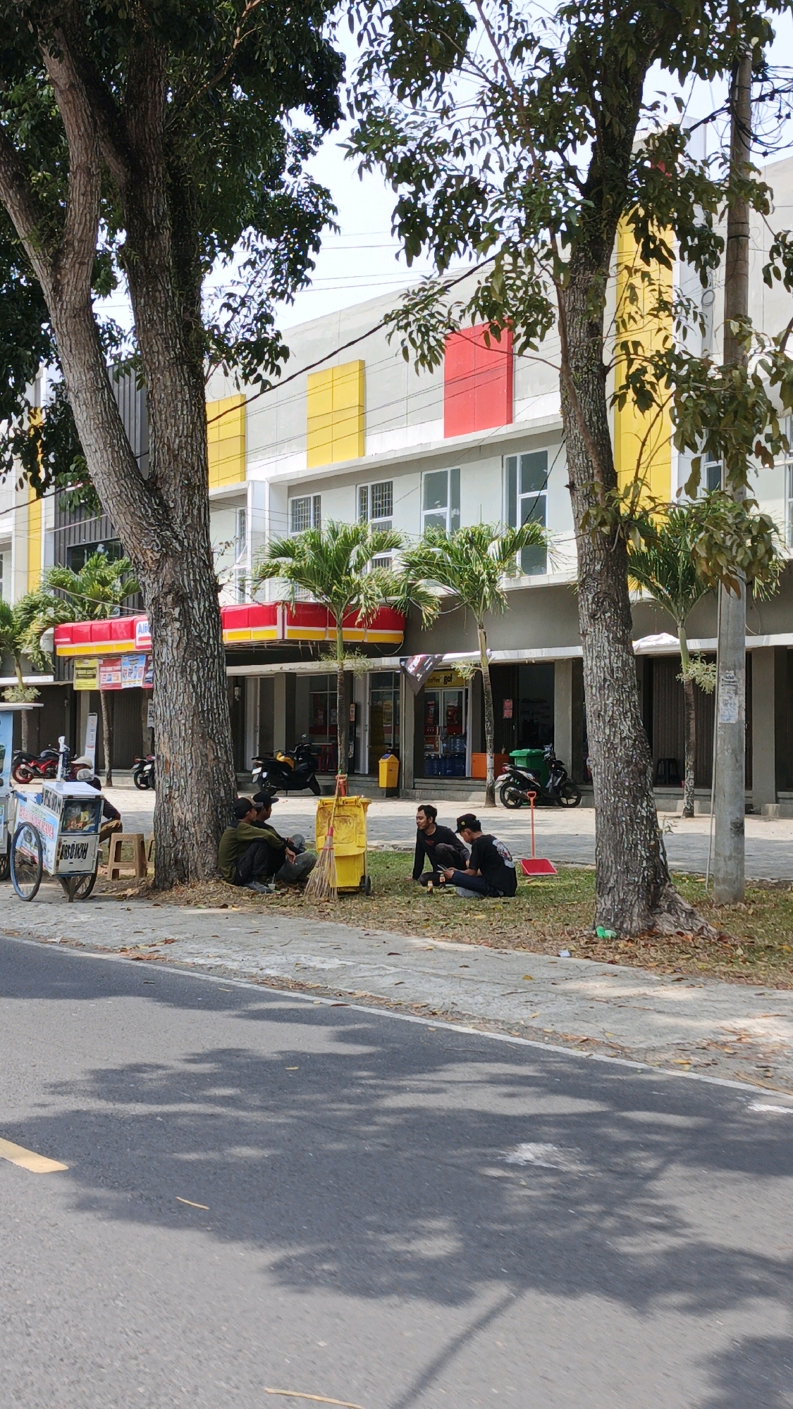 In the city of Desa Wonoharjo, Indonesia, four individuals are seated on the grass near the base of a large tree. Two individuals are positioned closer to the tree, one wearing a green shirt and the other a black shirt. Two more individuals are seated further to the right, both wearing black shirts. Between the groups of people, a yellow cleaning cart with a broom and dustpan is visible. To the left of the people, a mobile food cart with a white body and a blue stripe is parked on the sidewalk. In the background, a multi-story building with yellow and red accents on its facade is present. Several palm trees are planted in front of the building. Motorcycles are parked in front of the establishments on the ground floor. The scene appears to be on a sunny day, with tree shadows cast across the asphalt road in the foreground.