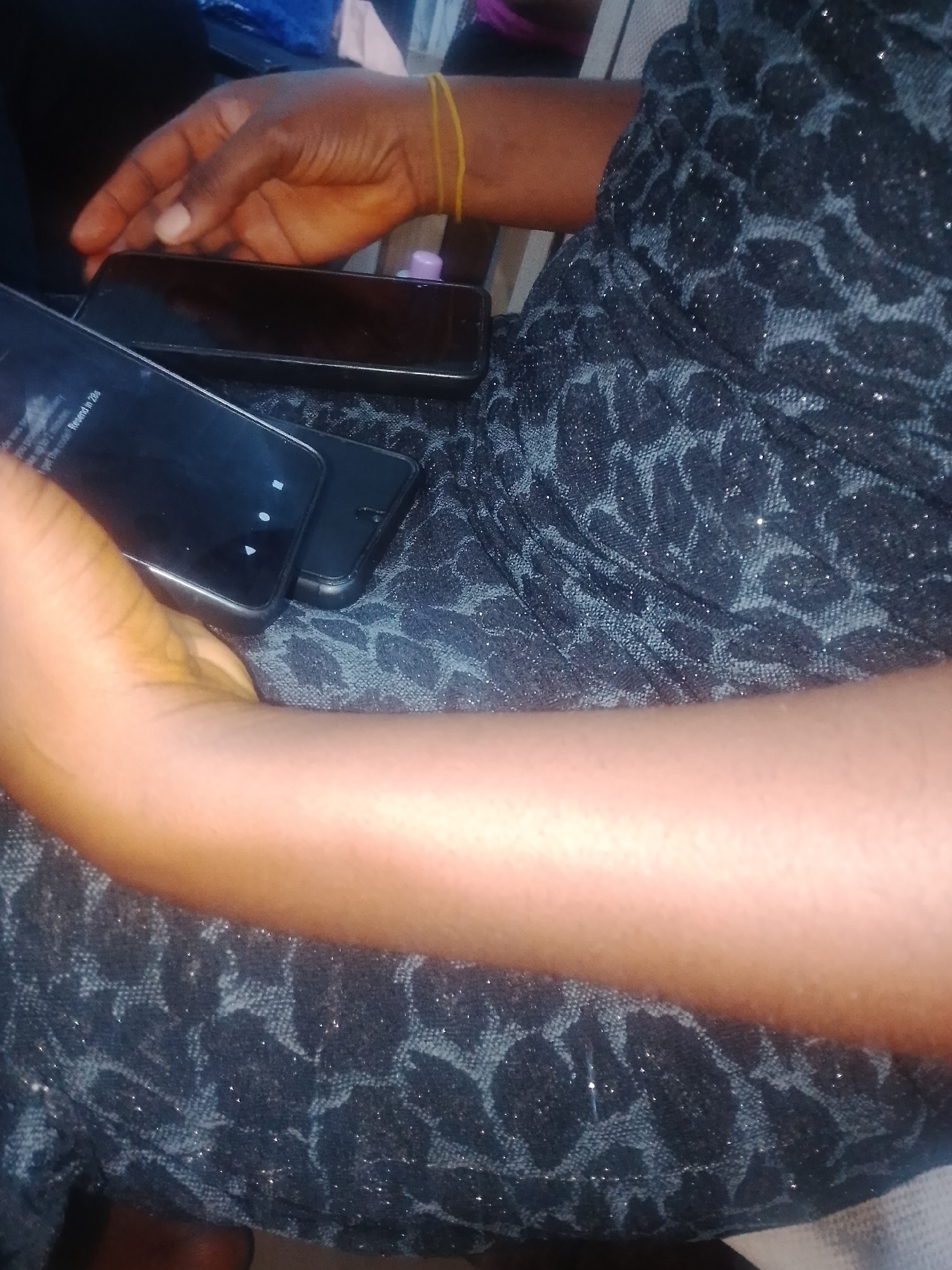 A person's arm and hand rest on top of two black smartphones. The person is wearing a black dress with a subtle leopard print pattern. A yellow elastic band is visible on the person's wrist. The smartphones are lying on a textured surface, possibly a table or lap. The background is dark and indistinct. The image appears to be taken indoors.