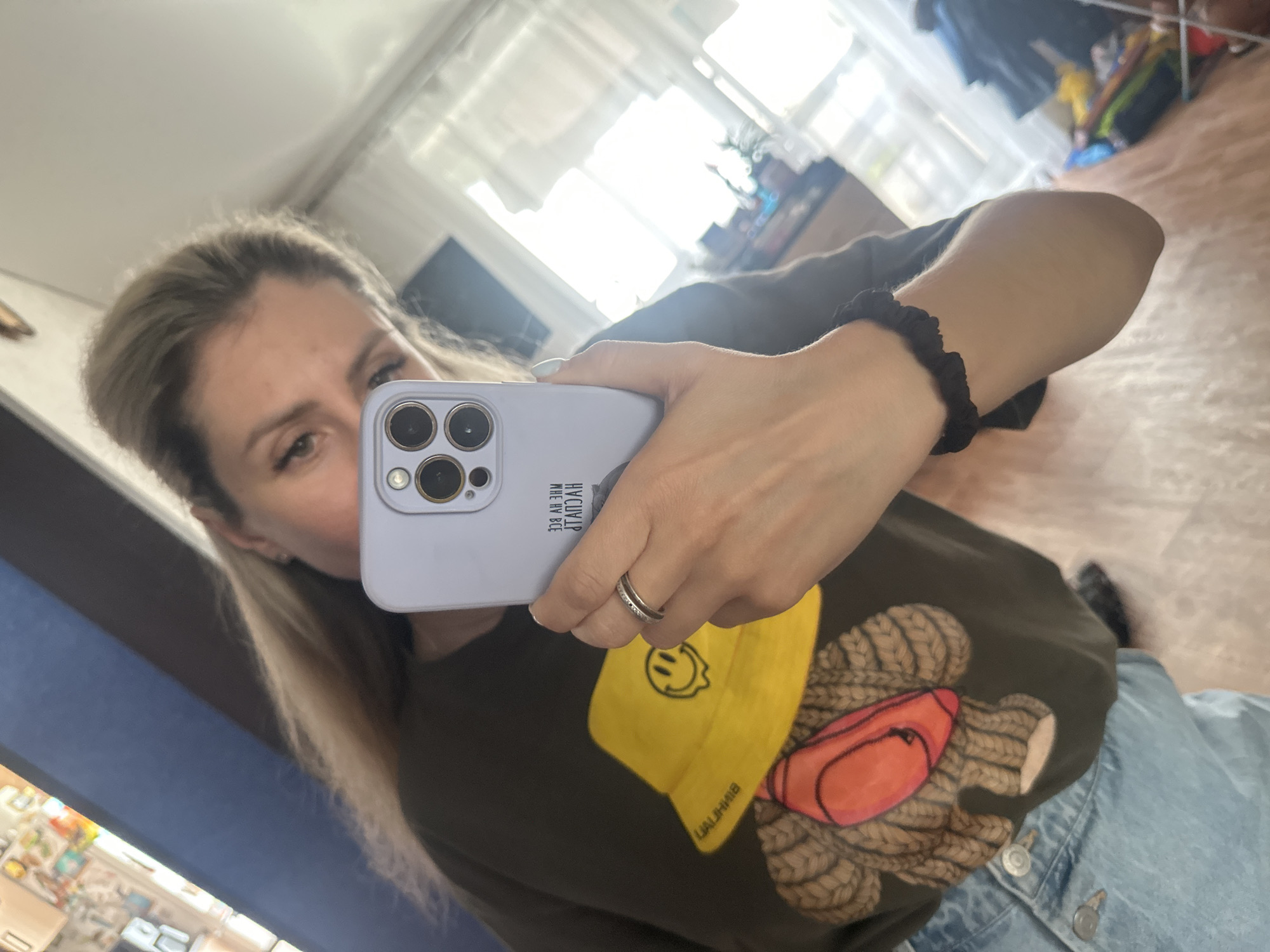 A woman takes a mirror selfie indoors. She is wearing a dark t-shirt with a graphic of a teddy bear wearing a yellow bucket hat and an orange fanny pack. She is also wearing light-wash denim jeans. She has a silver ring on her left ring finger and a black scrunchie on her left wrist. She is holding a light purple phone in her right hand, which is reflecting in the mirror. The background shows a room with a window, blinds, and furniture. The image appears to be taken in a residential setting in Novoaltaysk, Russia, given the context of other images from the same source.