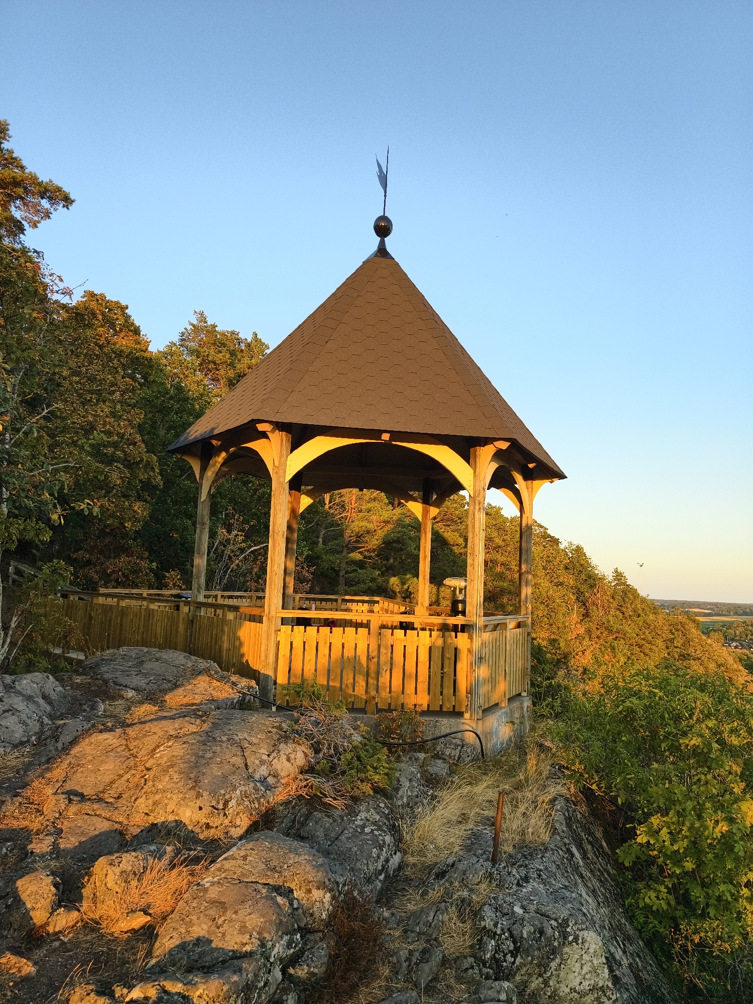 A wooden gazebo sits atop a rocky, tree-covered hill overlooking a distant landscape. The gazebo has a dark, shingled roof and an open wooden railing. Golden sunlight illuminates the structure and the surrounding foliage, suggesting it is either sunrise or sunset. A weathervane is visible on the peak of the gazebo's roof. The foreground consists of large, rough rocks with patches of dry grass. The background shows a dense forest and a hazy view of a valley or town. The image appears to be taken from a vantage point in Söderköping, Sweden, likely a nature reserve or a viewpoint. There are no people visible in the image.