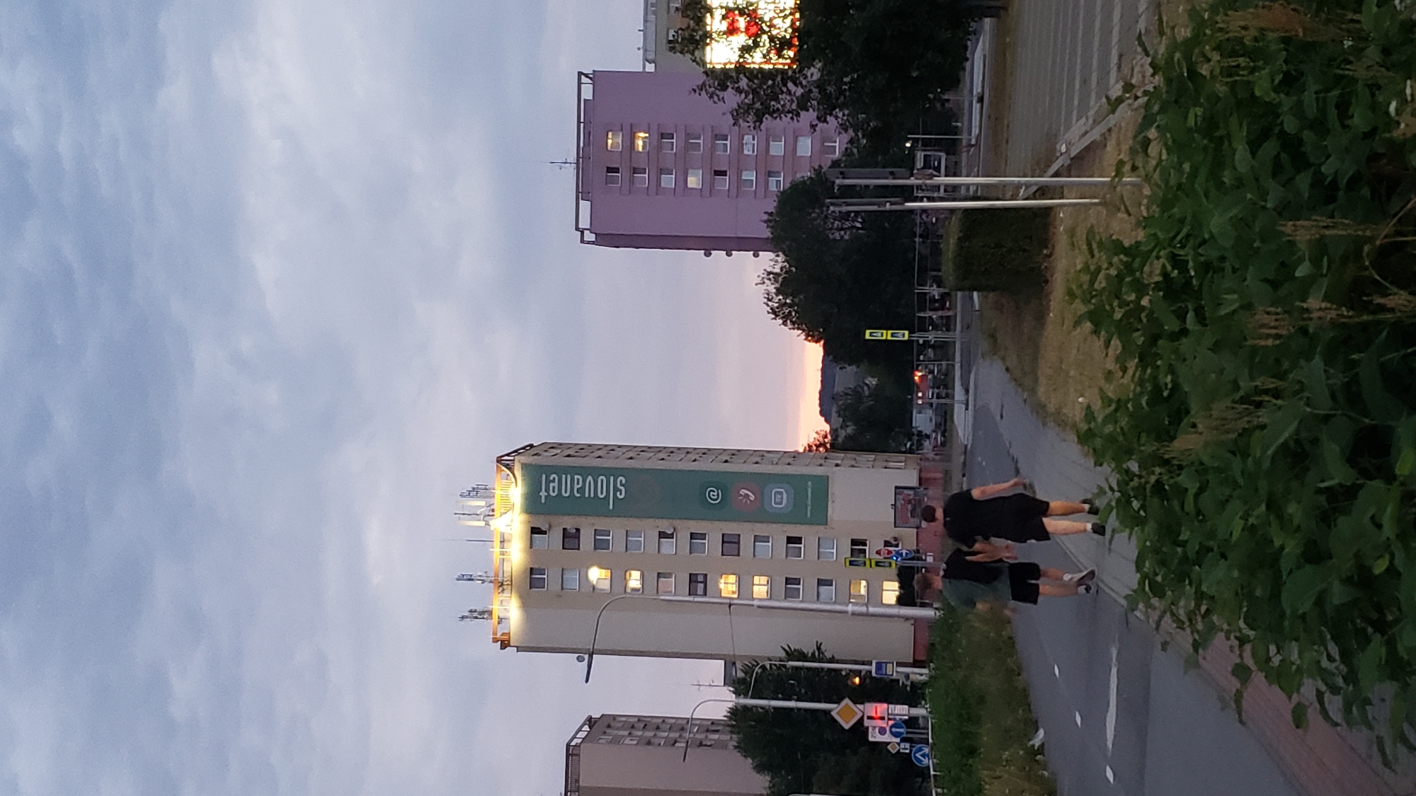 In Prešov, Slovakia, two young men walk away from the camera on a paved path, one in a green t-shirt and black shorts, the other in a black t-shirt and black shorts. They are walking towards two residential buildings. The building on the left is a tall, cream-colored apartment block with a large green banner displaying the text "slovaknet" and three circular icons. The building on the right is a shorter, lavender-colored apartment block. The sky above is overcast with a hint of sunset colors on the horizon. The foreground is filled with lush green foliage. The scene appears to be an urban residential area.