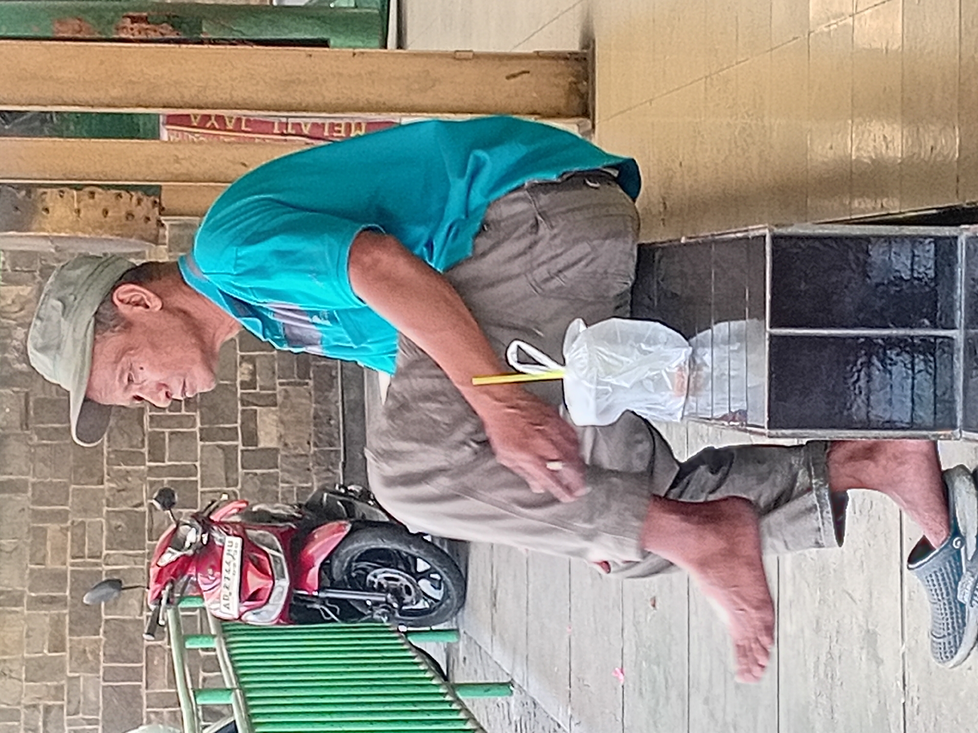 An elderly Asian man wearing a light green cap and a turquoise polo shirt sits on a black bench in Nganguk, Indonesia. He wears loose-fitting, tan pants and a single grey clog on his left foot. His right foot is bare and crossed over his left leg. He holds a cigarette in his right hand and a plastic bag with a yellow straw is to his left. A red and black scooter is parked behind him, to the left. The man is positioned in front of a stone wall with a green metal fence to his left. A vertical sign with red and yellow lettering is visible to his right. The ground appears to be tiled.