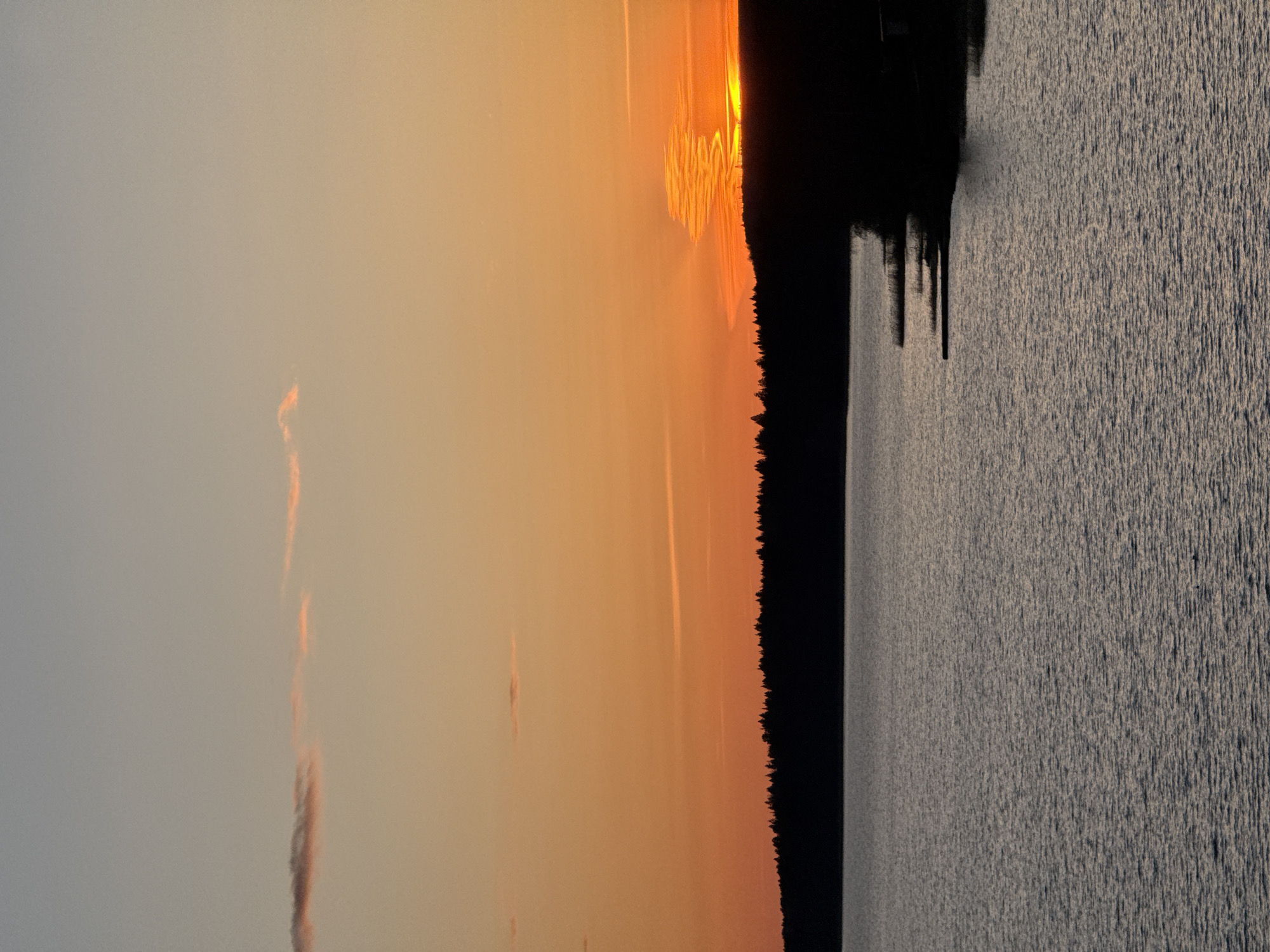 The image depicts a sunset over a body of water, likely a lake or reservoir, with a treeline silhouetted against the orange sky. The sun appears to be setting behind the trees. The water surface shows a rippled texture. While the scene is a natural landscape, the image does not contain any information directly pointing to the city of Votkinsk, Russia. Therefore, no details specific to that location can be inferred from the provided image.
