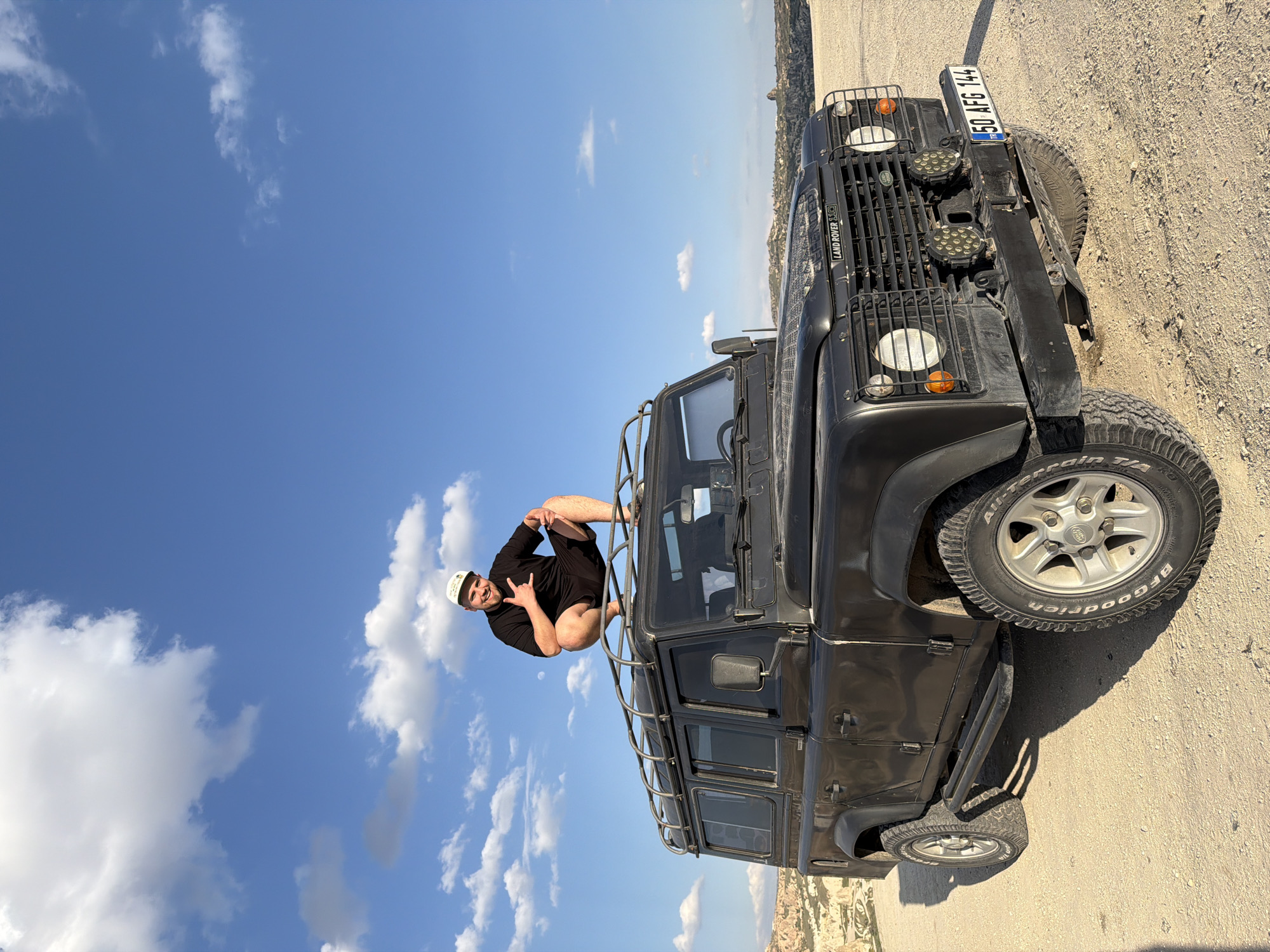 A bright, sunny outdoor scene shows a man posing playfully on the roof rack of a black Land Rover Defender, set against a backdrop of a clear blue sky with scattered white clouds. The image is taken on a dry, sandy, or dirt road, typical of an arid landscape.
The man, appearing to be a young adult with a beard, is wearing a black t-shirt or long-sleeved top, black shorts, and a white baseball cap with what appears to be a "PORSCHE" logo. He is squatting on the roof rack of the vehicle, making a "rock on" hand gesture with his left hand, and has a slight, cheerful smile on his face.
The vehicle is a rugged black Land Rover Defender 110, an off-road model, visible from the front three-quarter view. It features a distinctive front grille with visible text "LAND ROVER" and "DEFENDER 110," circular headlights, and two additional sets of circular LED lights mounted above the bumper. The license plate, visible on the front, reads "TR 50 AFG 144," indicating its origin in Turkey. The tires are BFGoodrich All-Terrain T/A, suggesting its readiness for various terrains.
In the distant background, faint rocky formations and valleys characteristic of the Göreme region in Cappadocia, Turkey, are visible under the bright daytime sun. The weather is clear and pleasant, with patches of cumulus clouds dotting the sky. The overall impression is one of adventure and enjoyment in a picturesque natural setting.