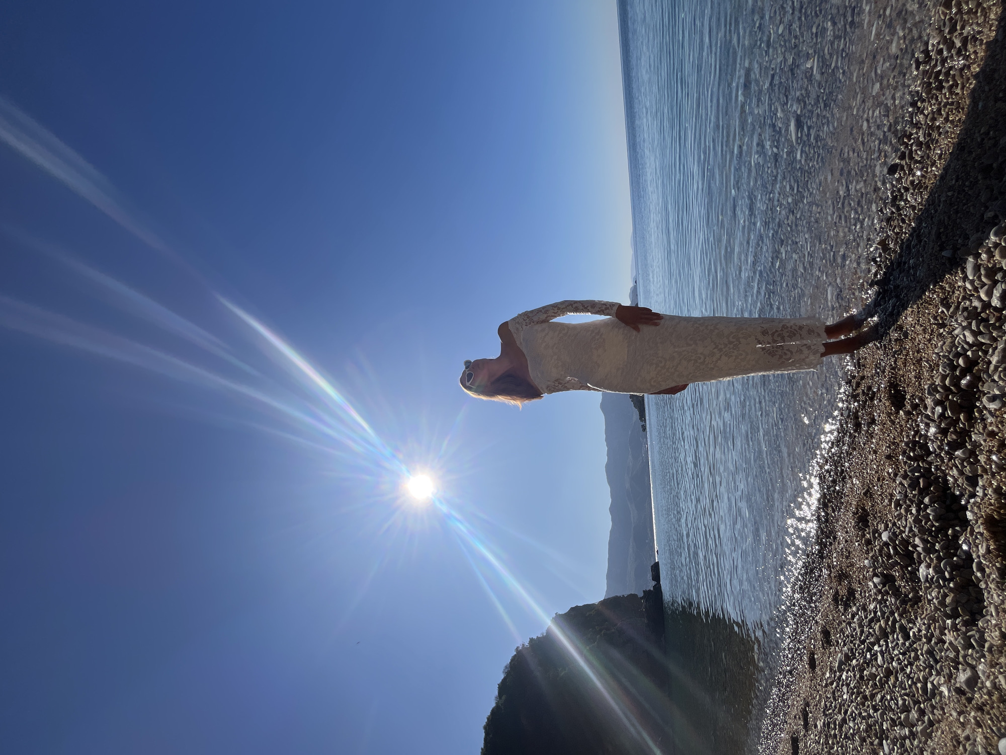 A woman in a white dress stands on a pebble beach, with her feet in the water. The sun is bright in the sky, and the water is calm. Mountains are visible in the distance. The scene is near Budva, Montenegro.