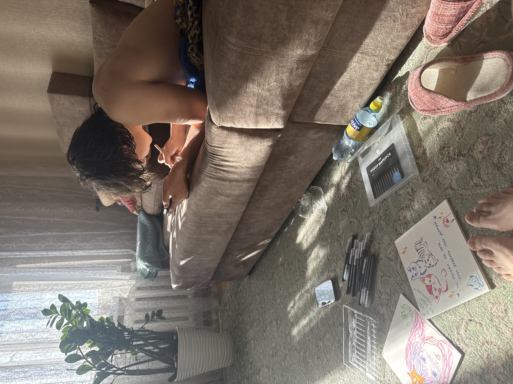 A person with wet dark hair is sitting on a beige sofa, drawing with markers on a piece of paper placed on a patterned carpet. A bottle of water, a package of markers, and slippers are nearby. A small mirror and several individual markers are also on the carpet. The person is holding a thin object, possibly a cigarette or drawing tool. A potted green plant is visible near a window covered with a sheer curtain. There is artwork present on the paper, including drawings of cats and an anime-style face. The OCR text suggests the package contains "markers" and phrases like "I want to take you home." The location is not specified.