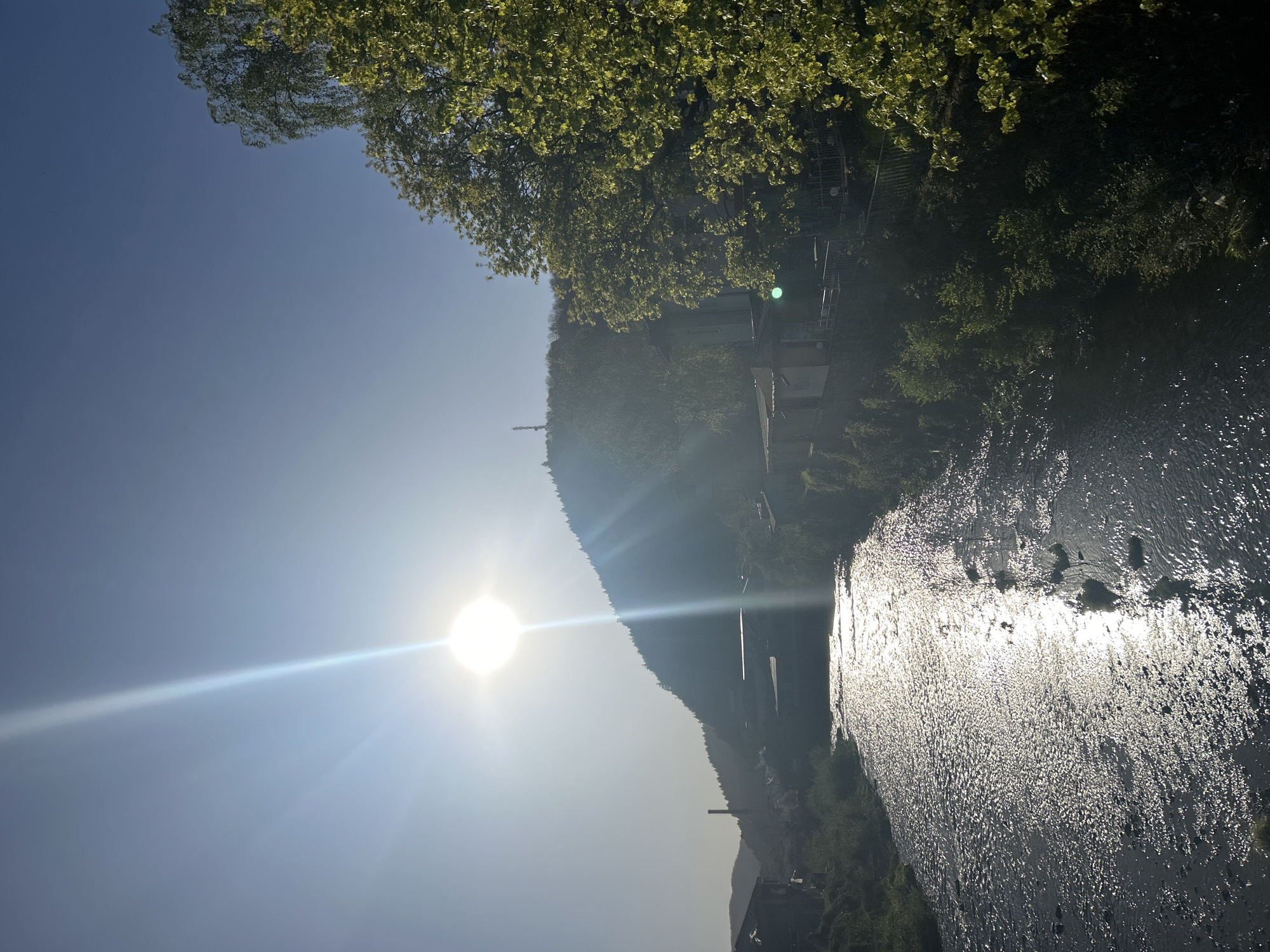 The image depicts the city of Zlatograd, Bulgaria. A river reflects the sunlight, and the opposite bank features dense vegetation interspersed with buildings and a hill. A prominent sun glare obscures some details. A tall, narrow structure stands on the hillside. The sky is clear.