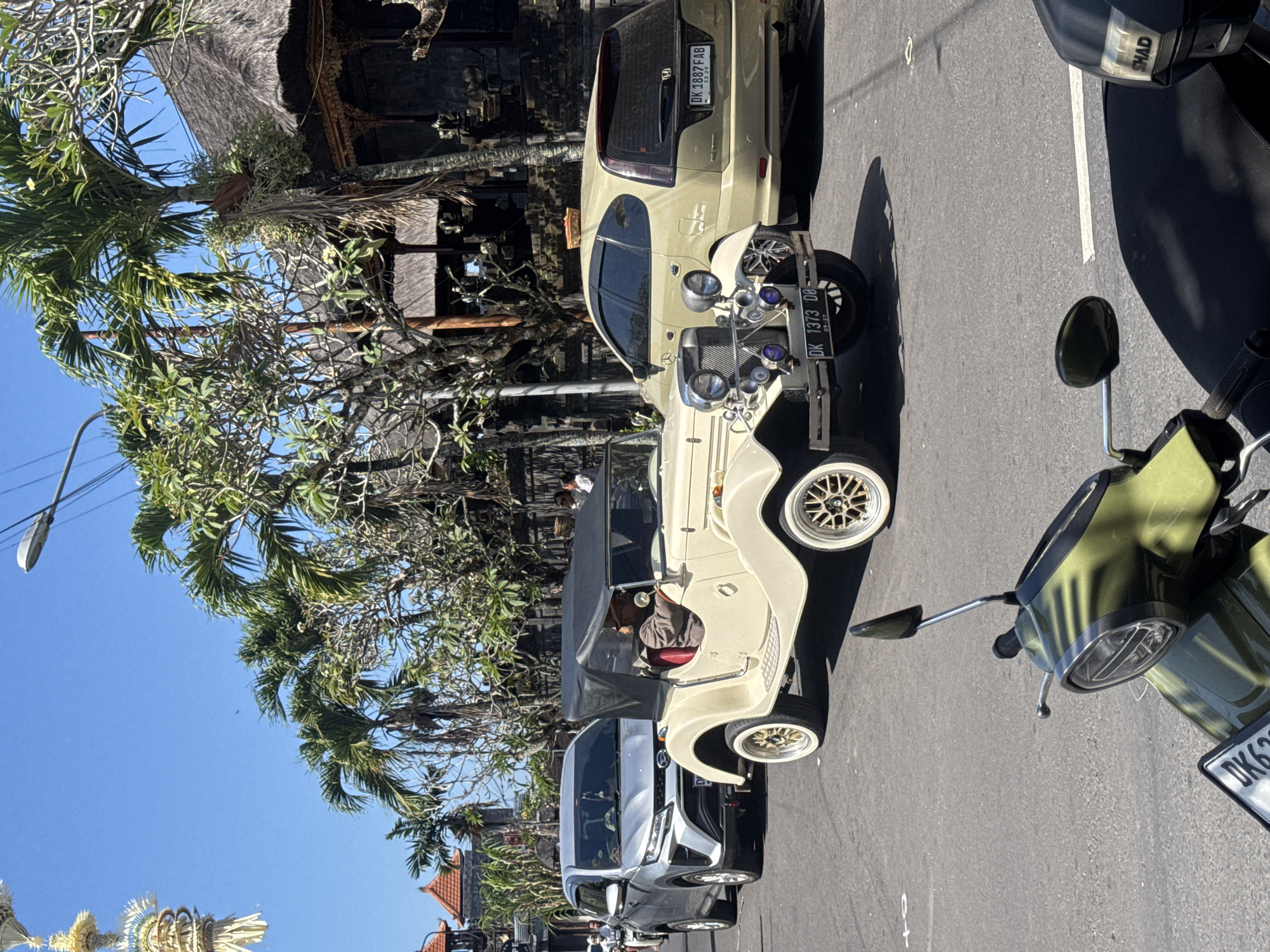 A vintage cream-colored convertible with a black soft top is driving on a street in Kuta Selatan, Indonesia. The car has a driver wearing a hat. A silver SUV is behind it, followed by a tan minivan with a license plate that reads "DK 1887 FAB". Palm trees and traditional Balinese architecture are visible in the background. A green scooter is parked in the foreground, partially obscuring the street. The sky is clear and blue, indicating a sunny day.