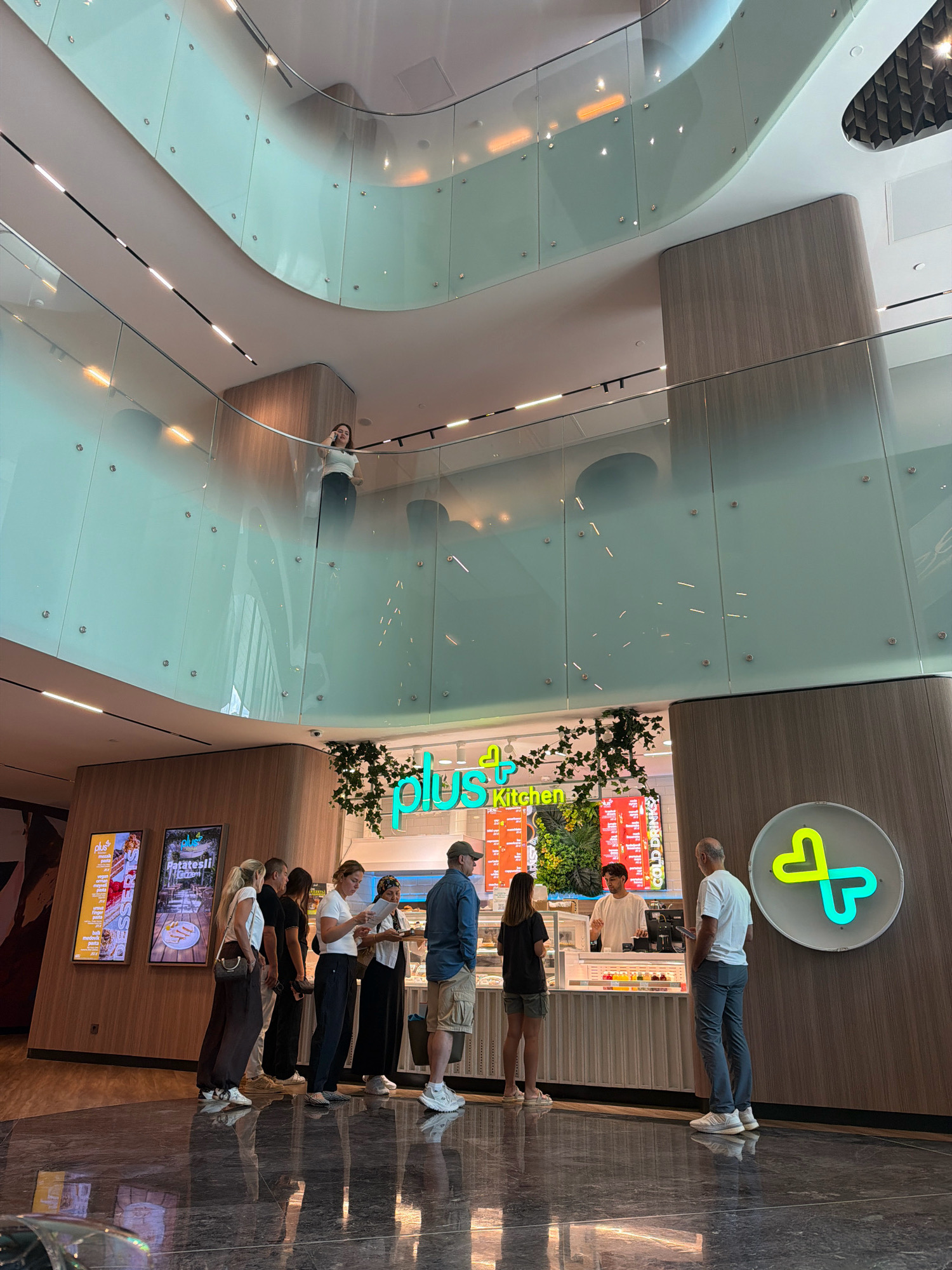The image depicts the interior of a modern establishment, likely a food vendor, located in Ataşehir, Turkey. The space features multiple levels with curved glass railings, creating an airy and contemporary aesthetic. A woman is visible on an upper level, standing near the railing and appearing to engage in a phone conversation. Below, at ground level, a queue of customers is forming in front of a counter labeled "plus+ Kitchen." The counter displays food items and a menu board with offerings such as "Desserts" and "Patatesli." Staff members are behind the counter, serving customers. A circular neon sign with a stylized heart logo is mounted on a wooden panel to the right of the counter. The floor is made of reflective dark marble. The overall environment suggests a bustling commercial area within a shopping mall or similar public space.