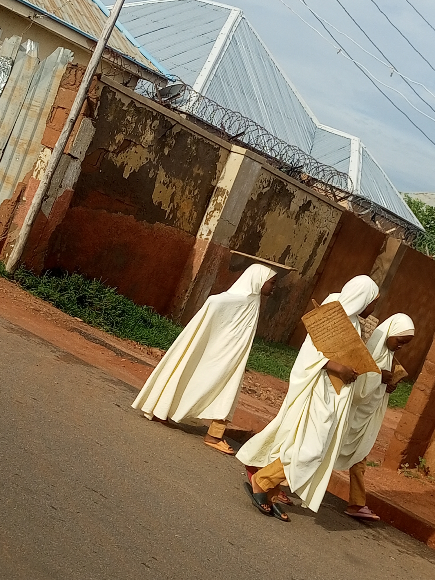 Three individuals wearing traditional white religious garments walk along a paved street. The person in the center is carrying a large, rectangular wooden tablet inscribed with text. The individual on the right is also holding a smaller inscribed tablet. They are walking on the right side of the street, with a textured, weathered wall bordering the street on the left. Above the wall, a corrugated metal roof is visible. The environment appears to be an outdoor urban setting in Gombe, Nigeria. The overall scene suggests a religious or educational context.